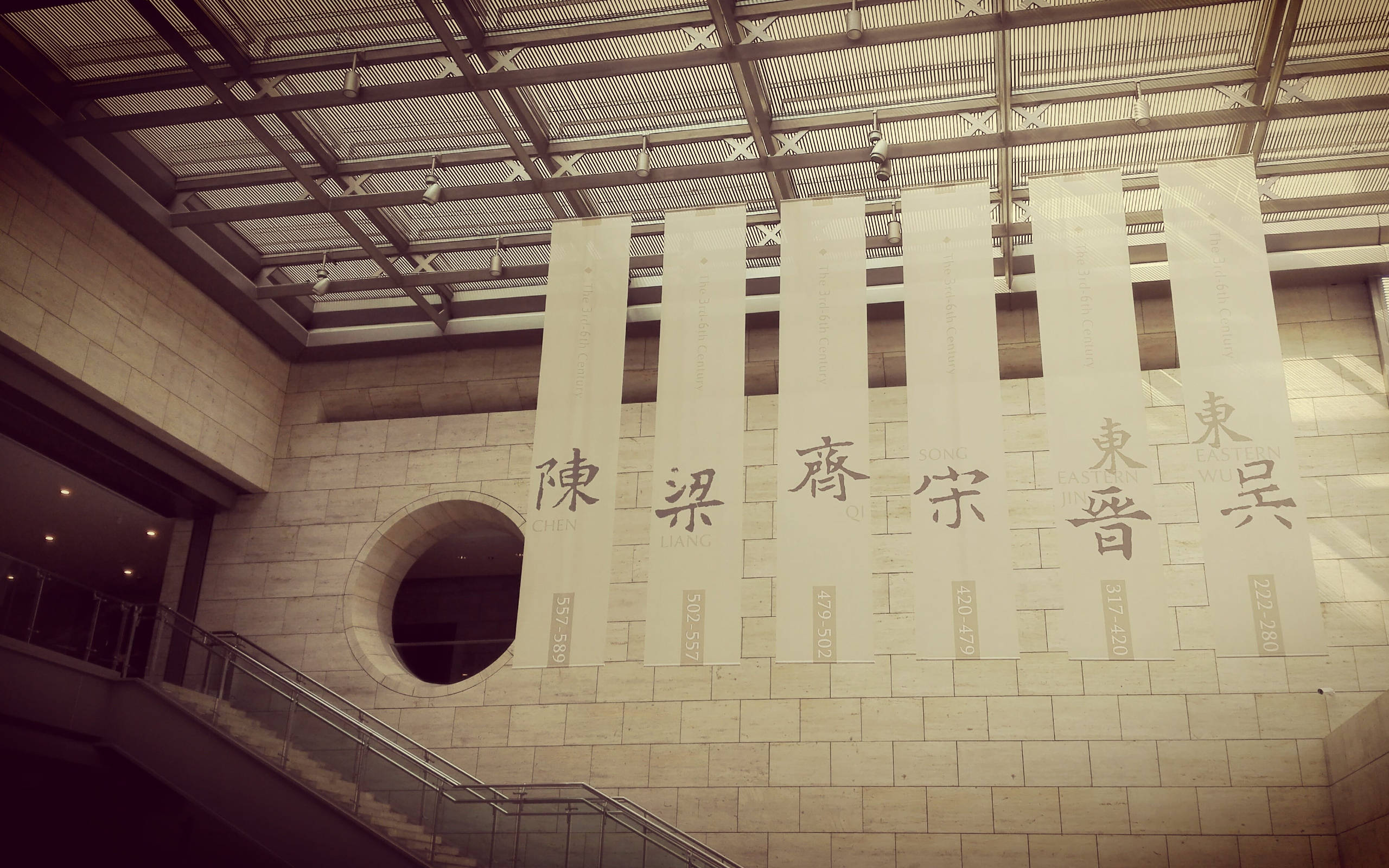南京六朝博物館 壁紙 — 現代博物館的棕褐色調室內景觀，掛有展示中文字元和日期的橫幅，背景為石牆和樓梯。