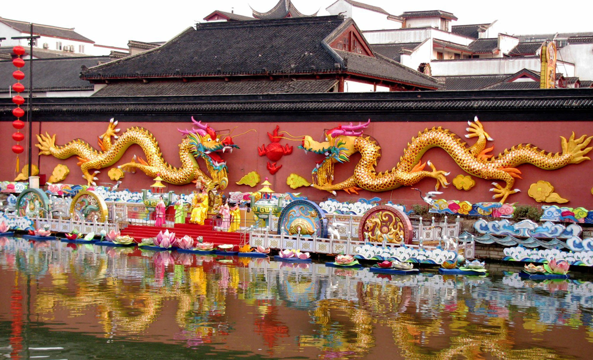 南京大神像屏 壁紙 — 鮮豔的金龍裝飾在紅牆上，下方是色彩繽紛的船隻和燈籠在平靜水面上倒影的節日水上巡遊。