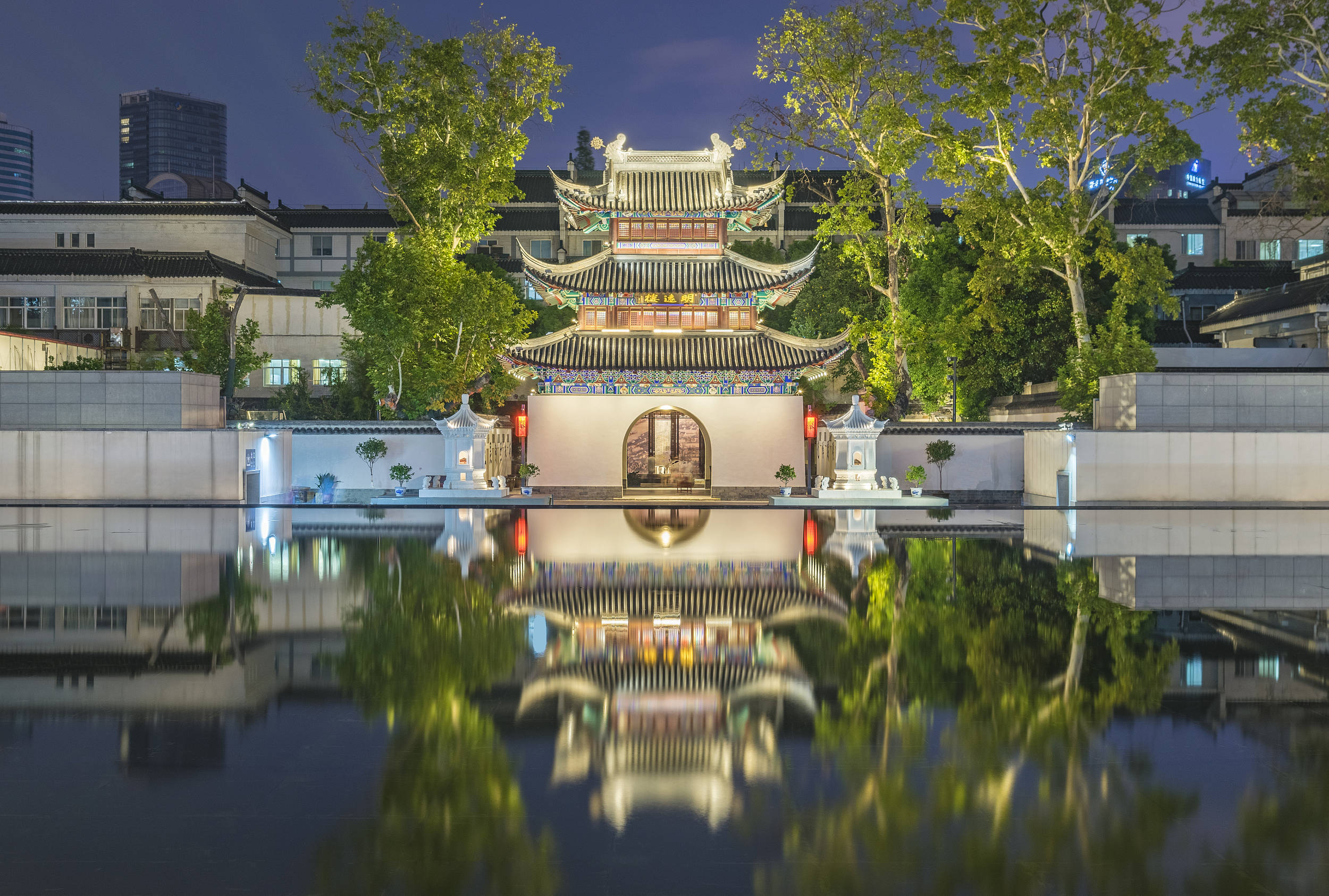 南京江南貢院博物館 壁紙 — 一座傳統中式亭台樓閣，飛簷翹角，在夜晚被美麗地照亮，其倒影完美地映照在平靜的水池中，四周環繞著鬱鬱蔥蔥的綠樹。