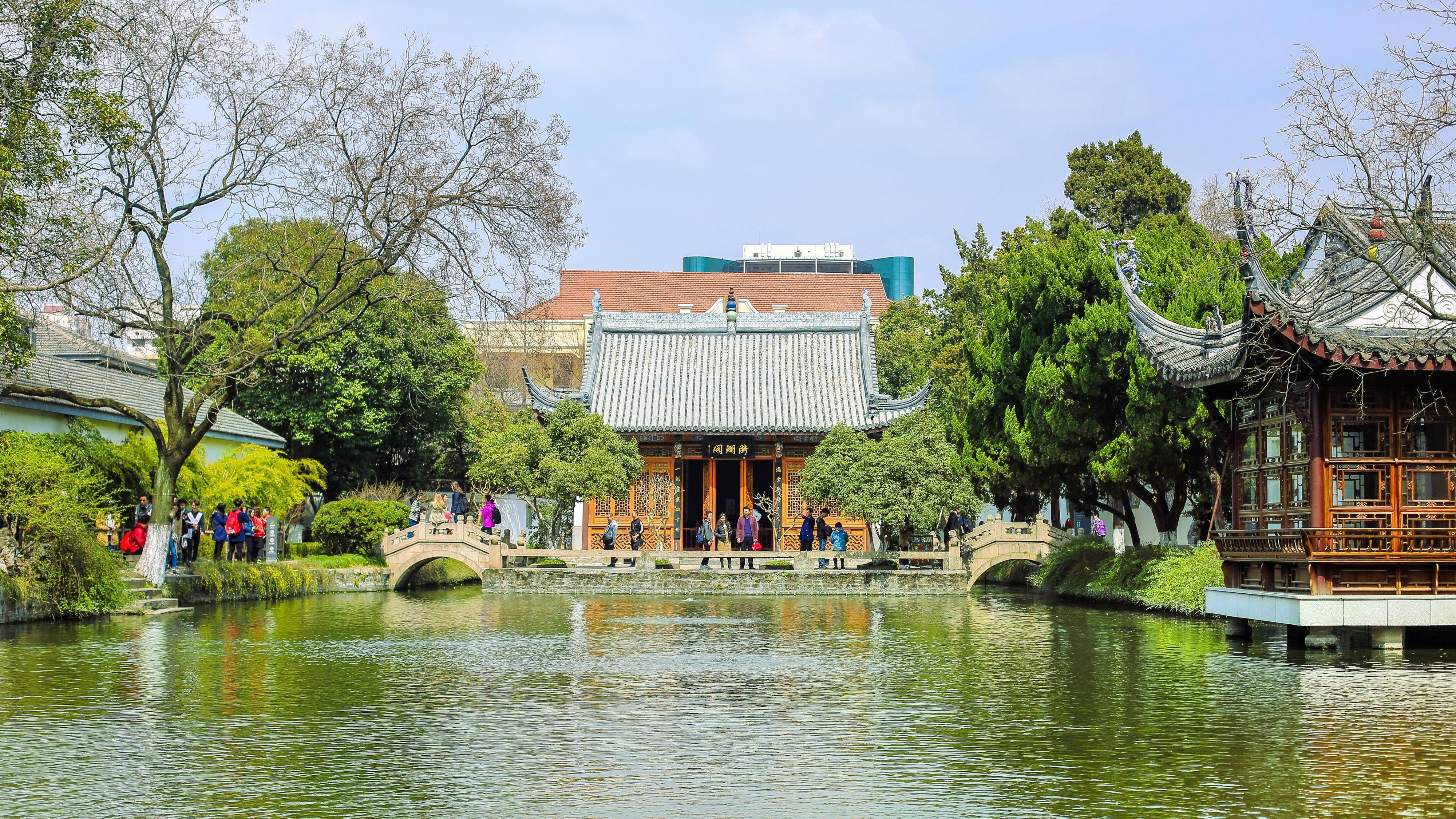 南京煦園 壁紙 — 傳統中式亭閣和拱橋倒映在平靜的水面上，四周環繞著茂密的綠植和遊客。