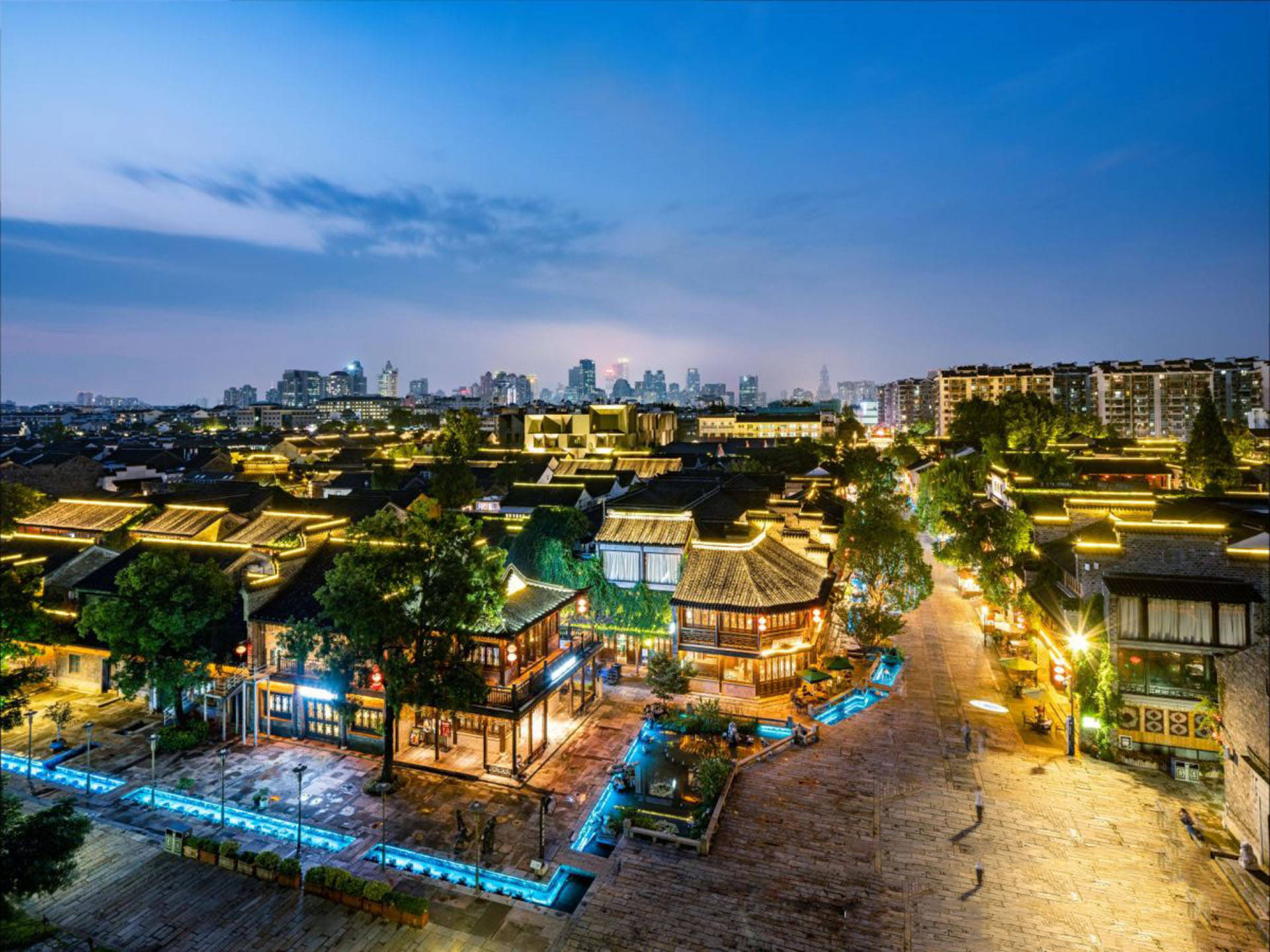 南京老門東古鎮傳統建築 壁紙 — 傳統瓦頂建築的鳥瞰圖，在暮色天空下散發溫暖燈光，遠方有現代摩天大樓。