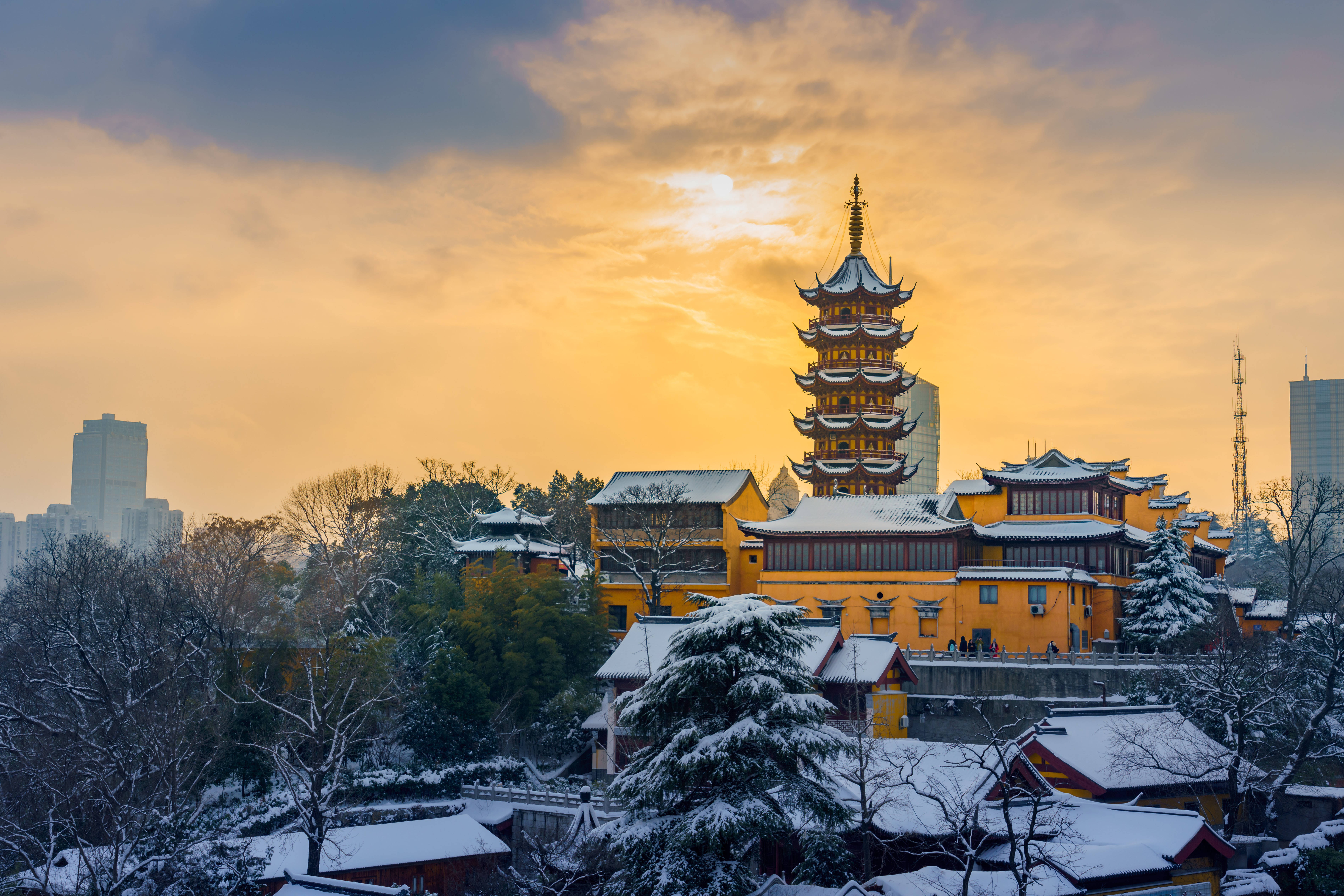 南京雞鳴寺雪景 壁紙 — 一個被雪覆蓋的傳統寶塔和寺廟建築群，在金色夕陽天空下溫暖地發光，背景是現代摩天大樓。