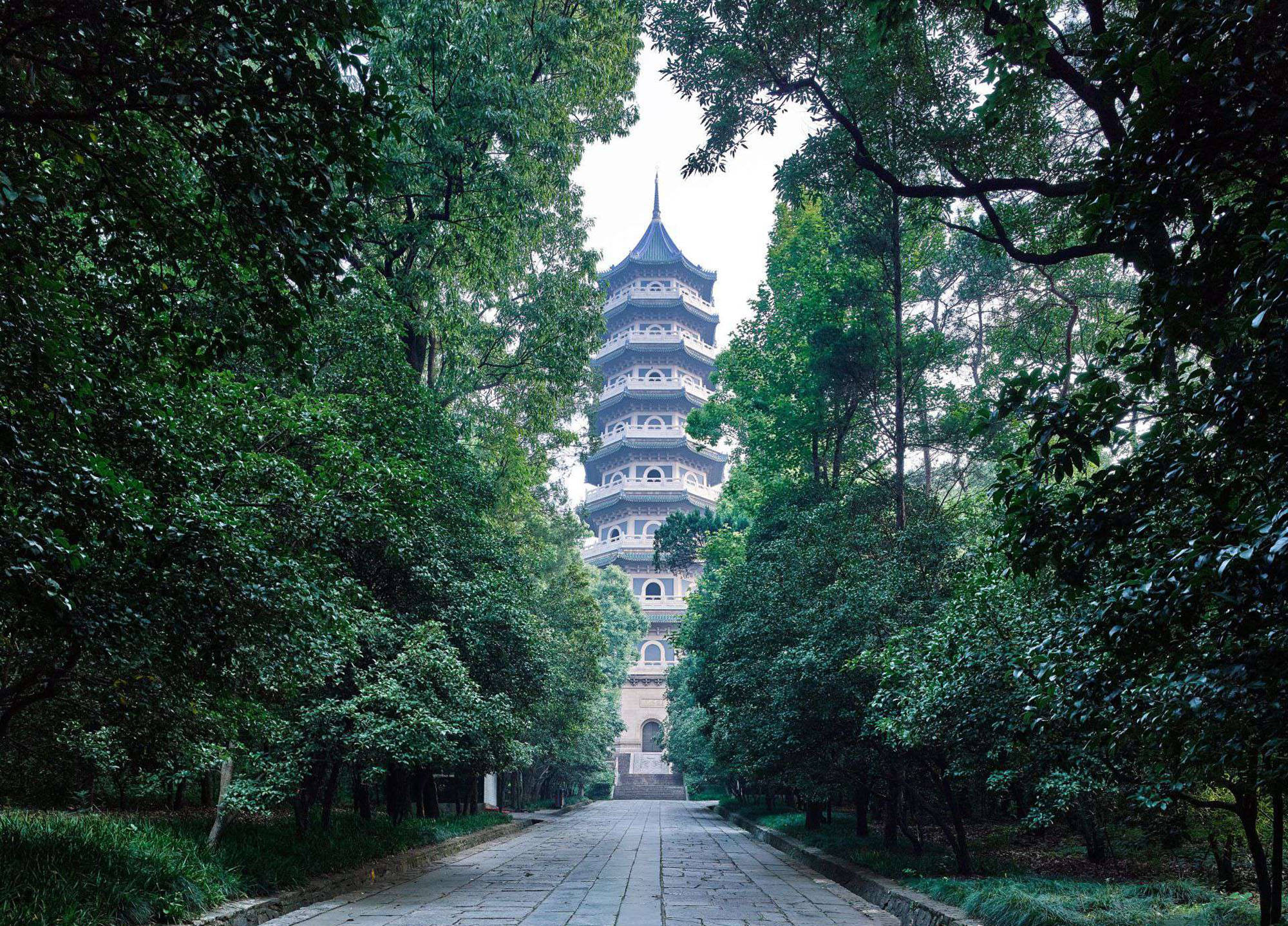 南京靈谷寺寶塔 壁紙 — 一條寧靜的石徑蜿蜒穿過茂密的綠樹，通往一座傳統的多層寶塔，天空多雲。