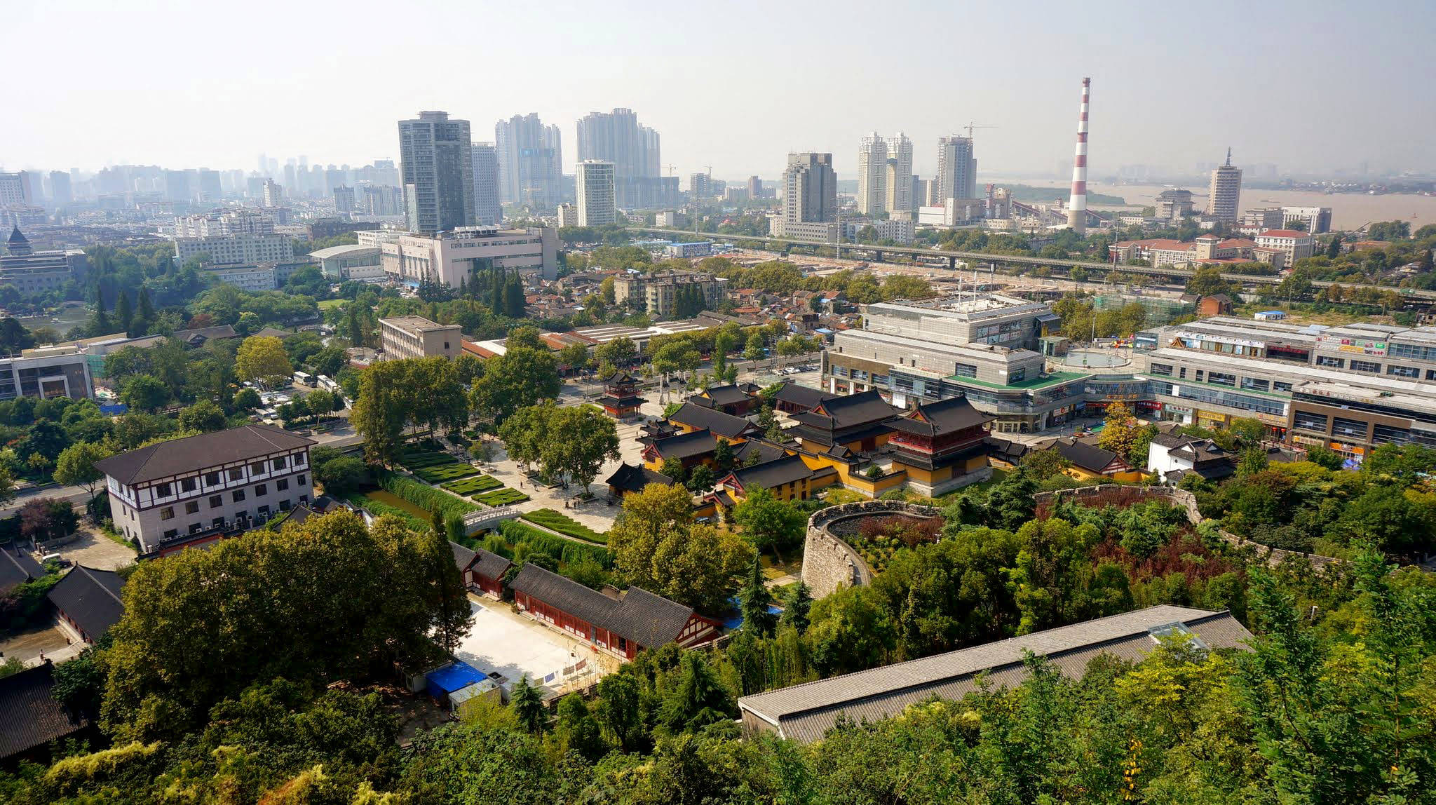 南京鼓樓全景 壁紙 — 在朦朧的日光下，現代城市天際線中鬱鬱蔥蔥的綠樹環繞著的傳統中式寺廟的鳥瞰圖。