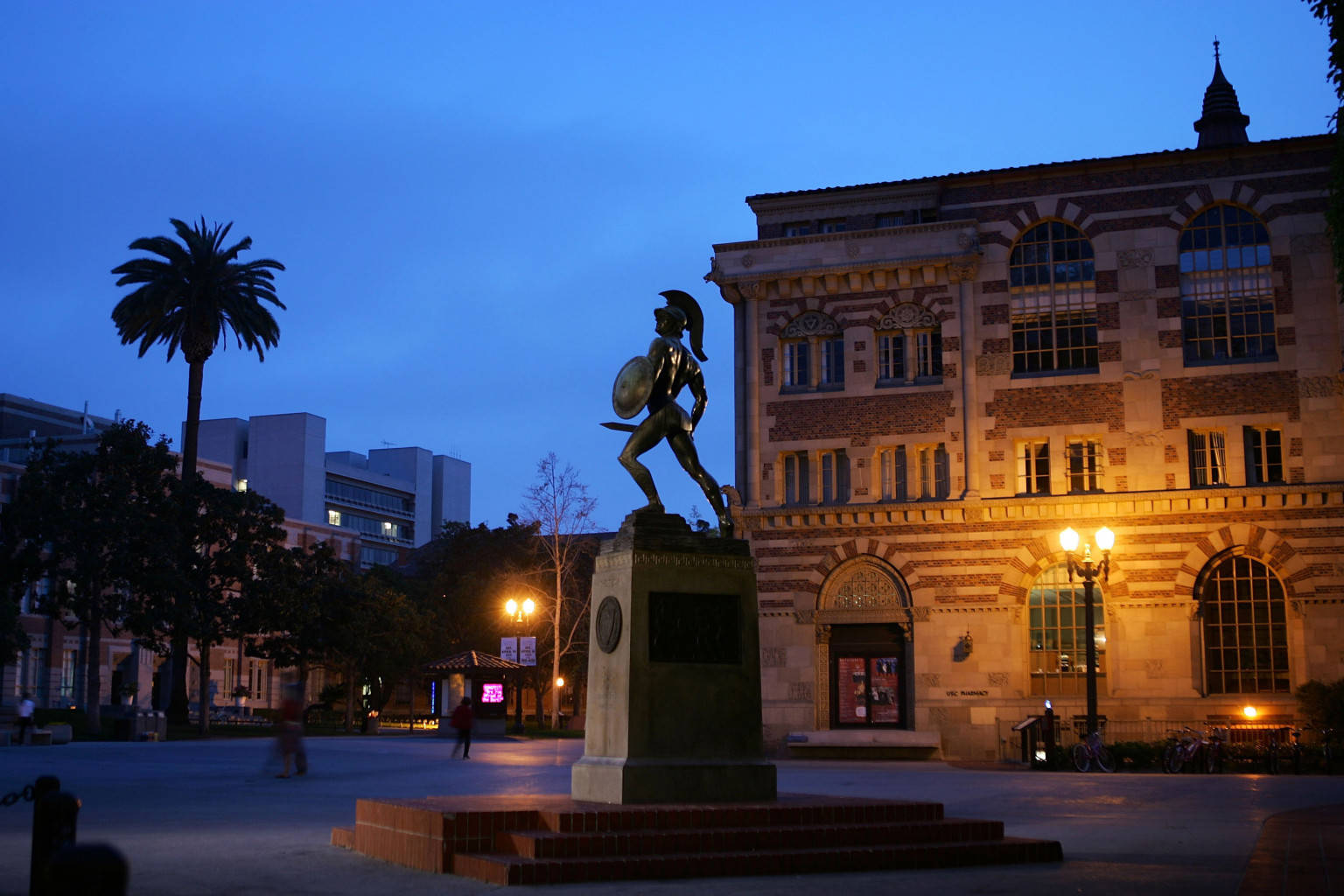 南加州大學湯米特洛伊雕像 壁紙 — 一座青銅斯巴達戰士雕像在黃昏時分被照亮，矗立在一座帶有拱形窗戶和溫暖燈光的宏偉石建築前。