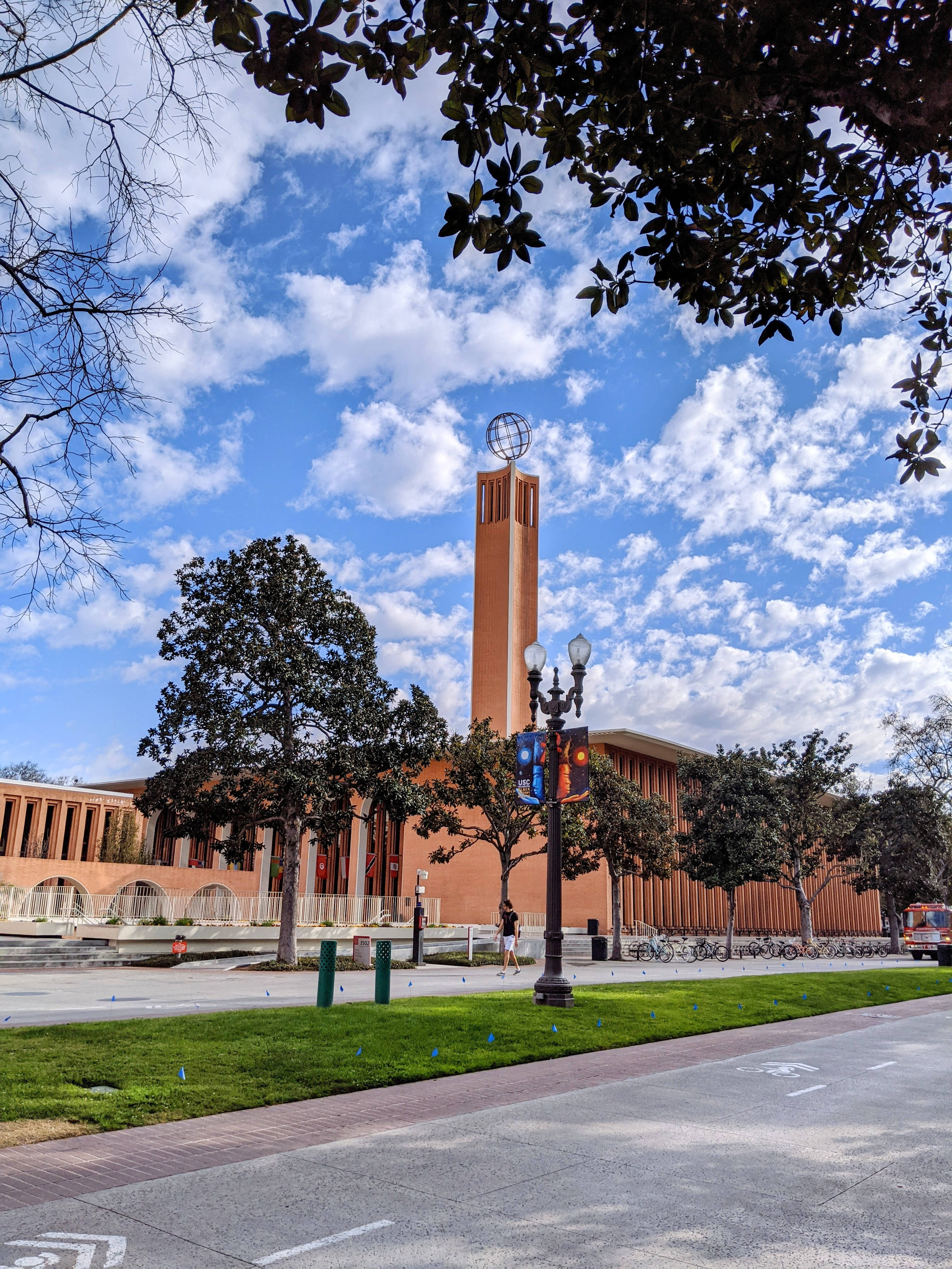 南加州大學馮·克萊因斯米德中心 壁紙 — 一座高大的赤陶塔頂端有一個地球儀，在樹枝環繞的明亮藍天和蓬鬆白雲襯托下聳立。