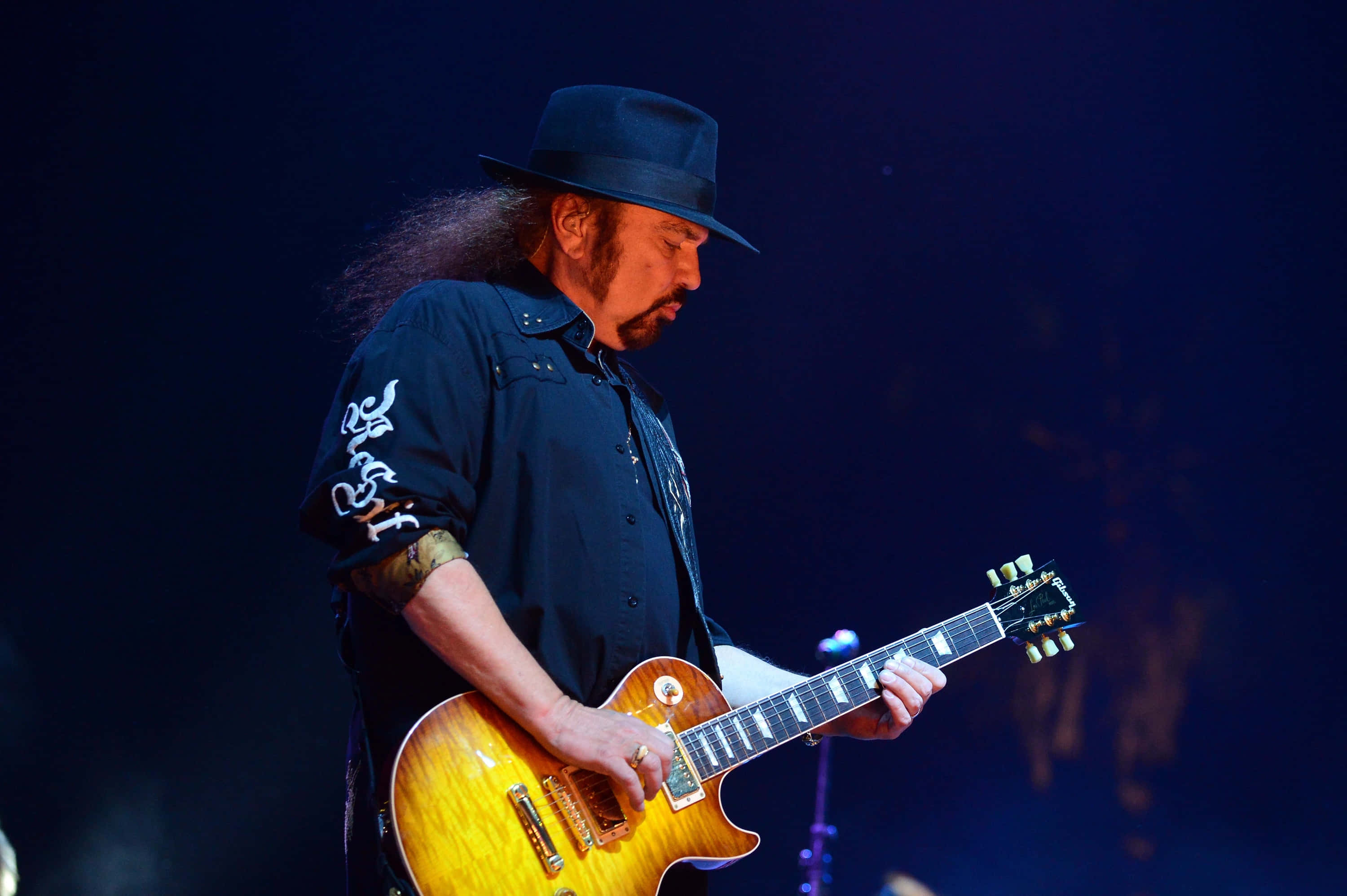 南方搖滾樂隊 Lynyrd Skynyrd 的創始成員兼主音吉他手 Gary Rossington 壁紙 — 一位長髮戴黑帽的音樂家在舞台燈光下彈奏一把旭日紋電結他。