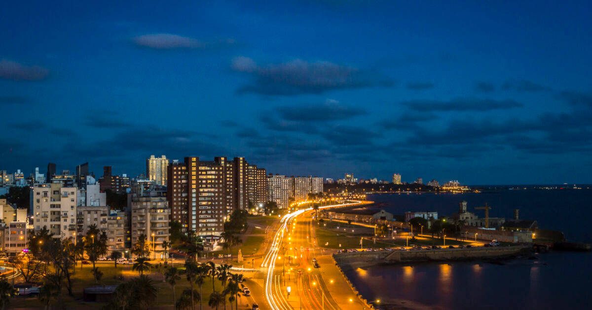 南美洲 烏拉圭城市夜景 壁紙 — 黃昏時分充滿活力的城市景觀，發光的街燈沿著海濱道路延伸，映襯著深藍色的天空。