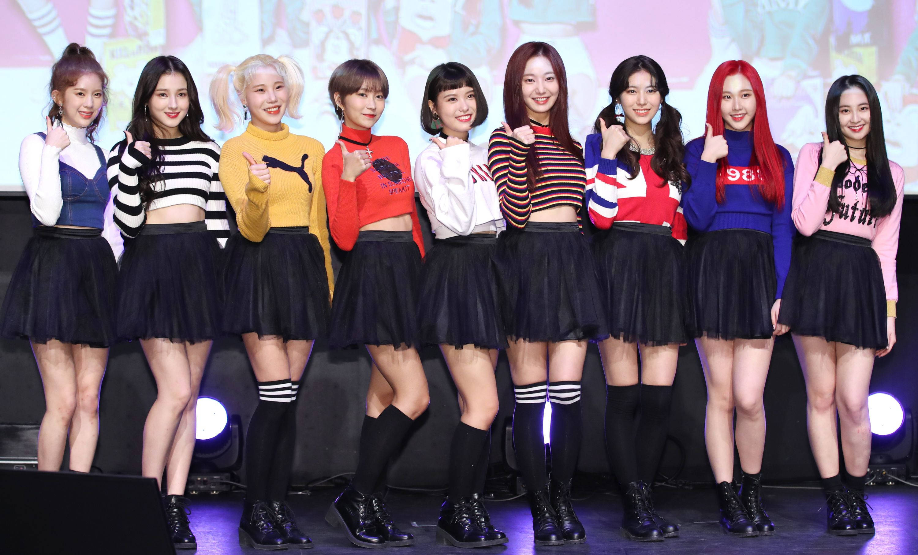 南西 Momoland 高畫質與全體成員 壁紙 — 九位微笑的女性偶像穿著色彩繽紛的露臍上衣和黑色裙子，在明亮的舞台燈光下自信地擺姿勢，身後有明亮的聚光燈。