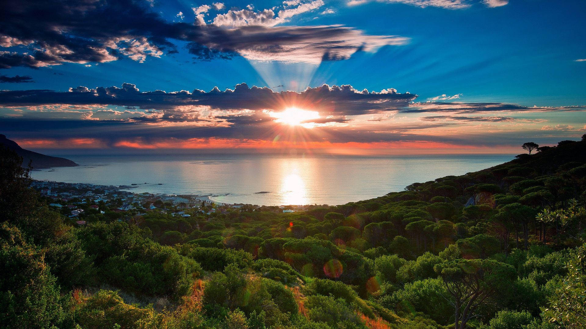 南非日落海景 壁紙 — 陽光穿透雲層照射在海面上，金色的光芒在水面上反射，周圍環繞著鬱鬱蔥蔥的綠色山丘和遠處的海岸小鎮。