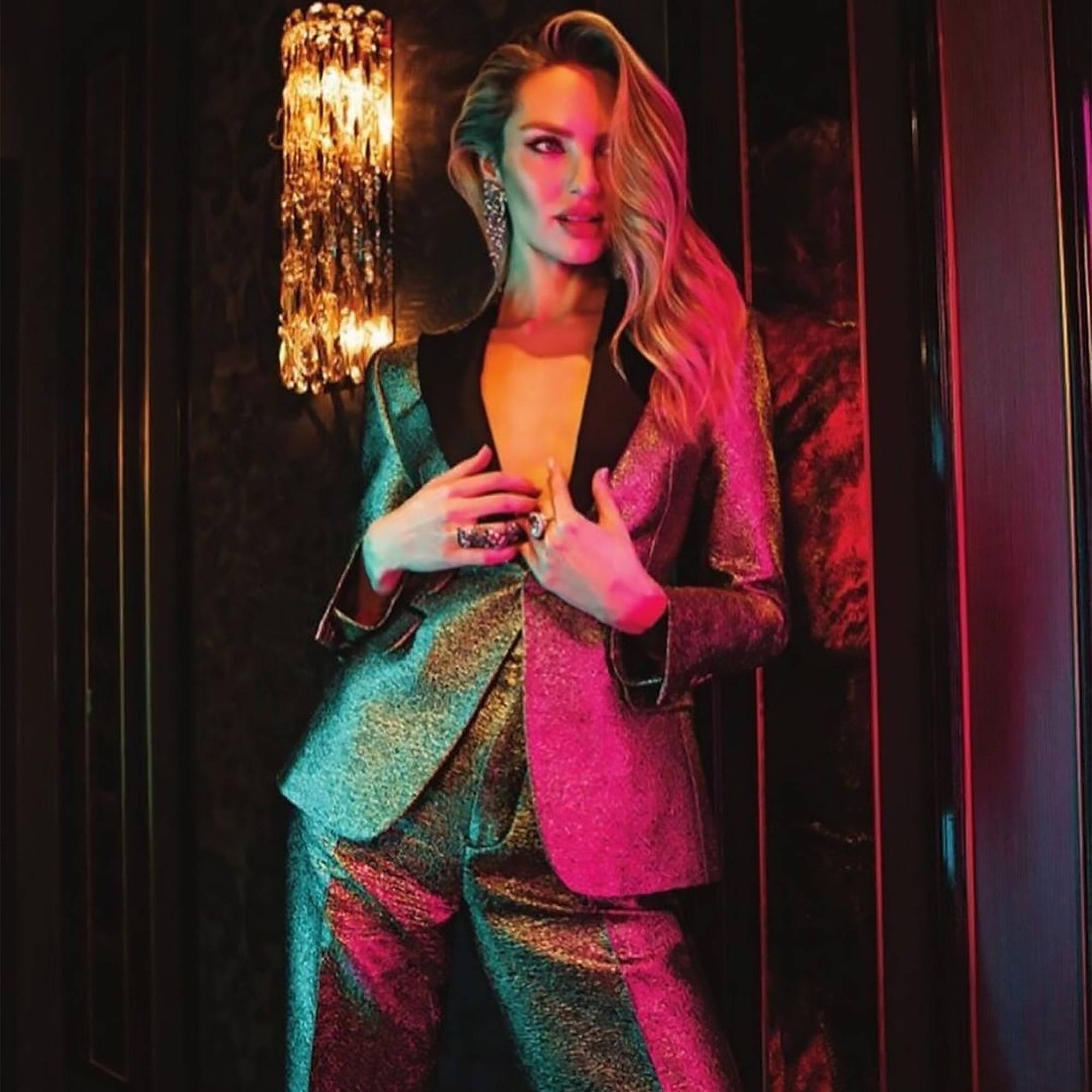南非超模 Candice Swanepoel 登上雜誌封面 壁紙 — 一位身穿閃亮雙色套裝的迷人女性，在溫暖的吊燈燈光下，以深色天鵝絨牆壁為背景擺出姿勢。