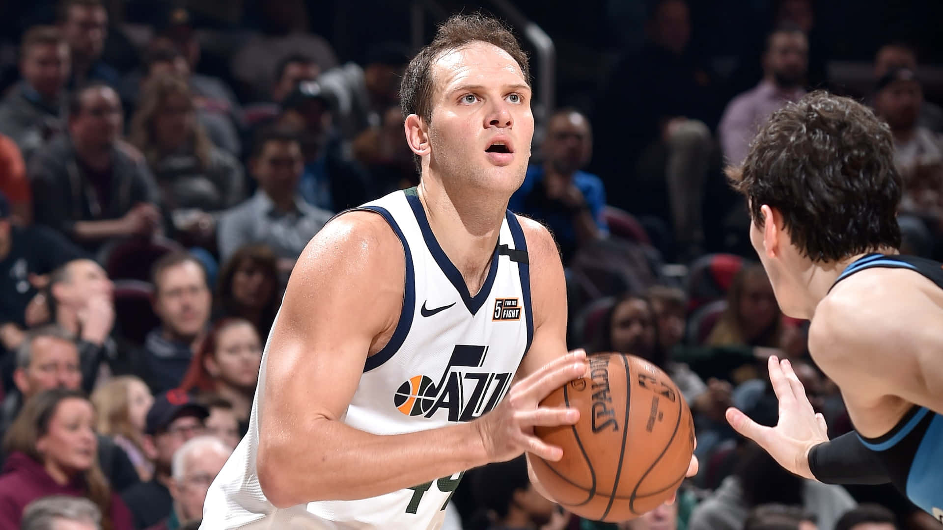 博揚·博格達諾維奇 NBA 聯盟 爵士隊 壁紙 — 一名身穿白色爵士隊球衣的籃球運動員在擠滿觀眾的場館中運球，同時被對手防守。