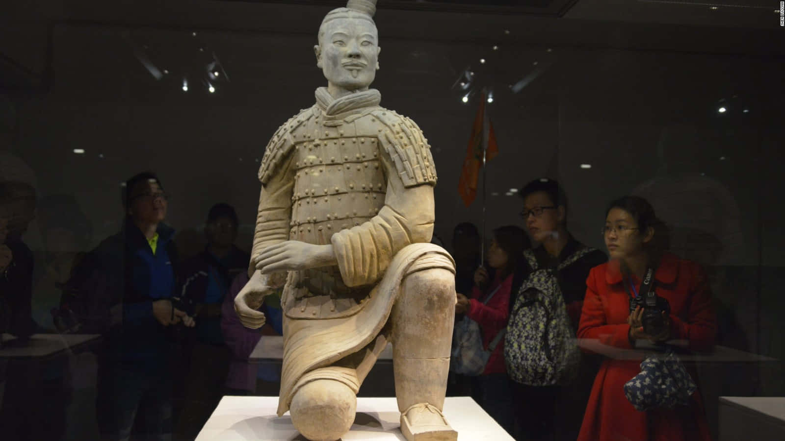 博物館內兵馬俑的壯麗展覽 壁紙 — 博物館展覽中一個跪著的兵馬俑雕像，背景有參觀者在觀察。