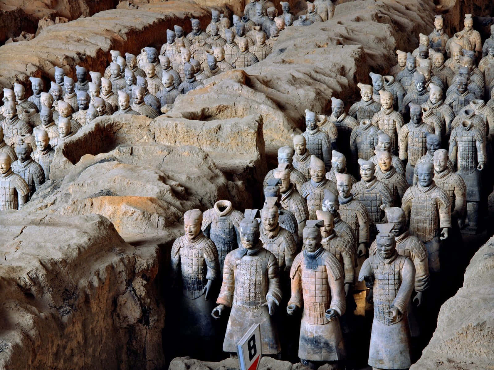博物館內的兵馬俑葬禮藝術 壁紙 — 一排排古老的兵馬俑站在泥土的發掘坑中，帶有詳細的盔甲和多樣的表情。