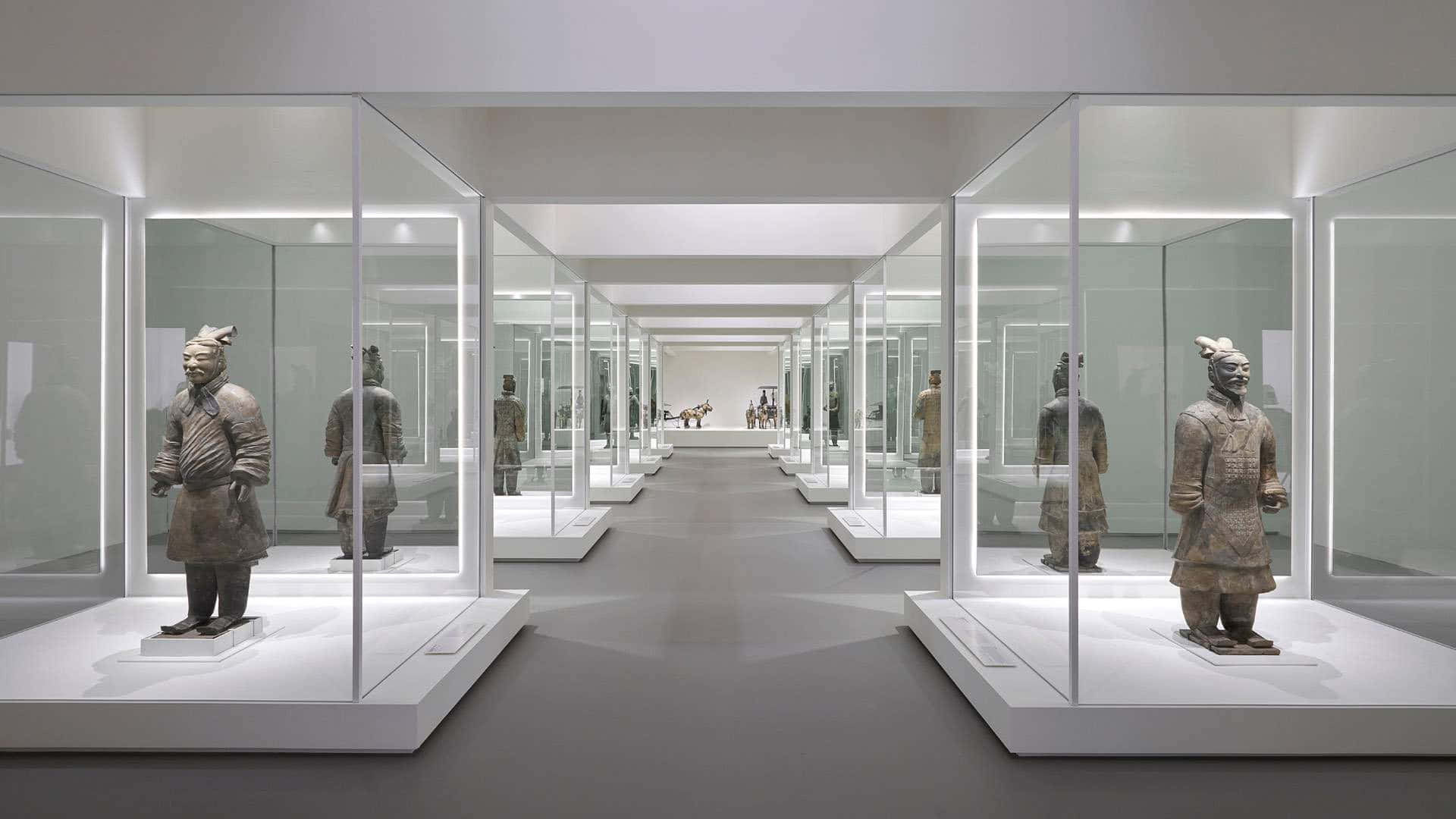 博物館內秦俑雕塑群展示 壁紙 — 一條長廊，兩旁玻璃陳列櫃在明亮白光下展示古代兵馬俑。