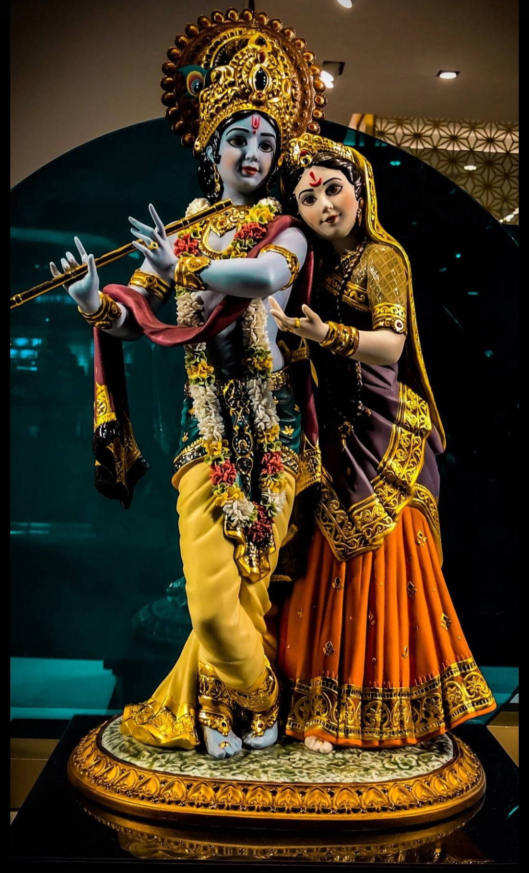 krishna 3d 桌布 壁紙 — 博物館展覽中，一尊色彩鮮豔的克里希納和拉達雕像，身著華麗的金色和橙色服飾，正在吹奏長笛。