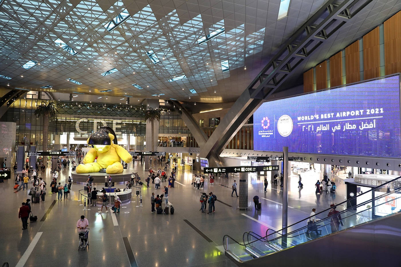卡塔爾哈馬德國際機場壯麗夜景 壁紙 — 一個繁忙的現代機場客運大樓，有一個巨大的黃色吉祥物雕像和一個顯示「World's Best Airport 2021」的大屏幕。