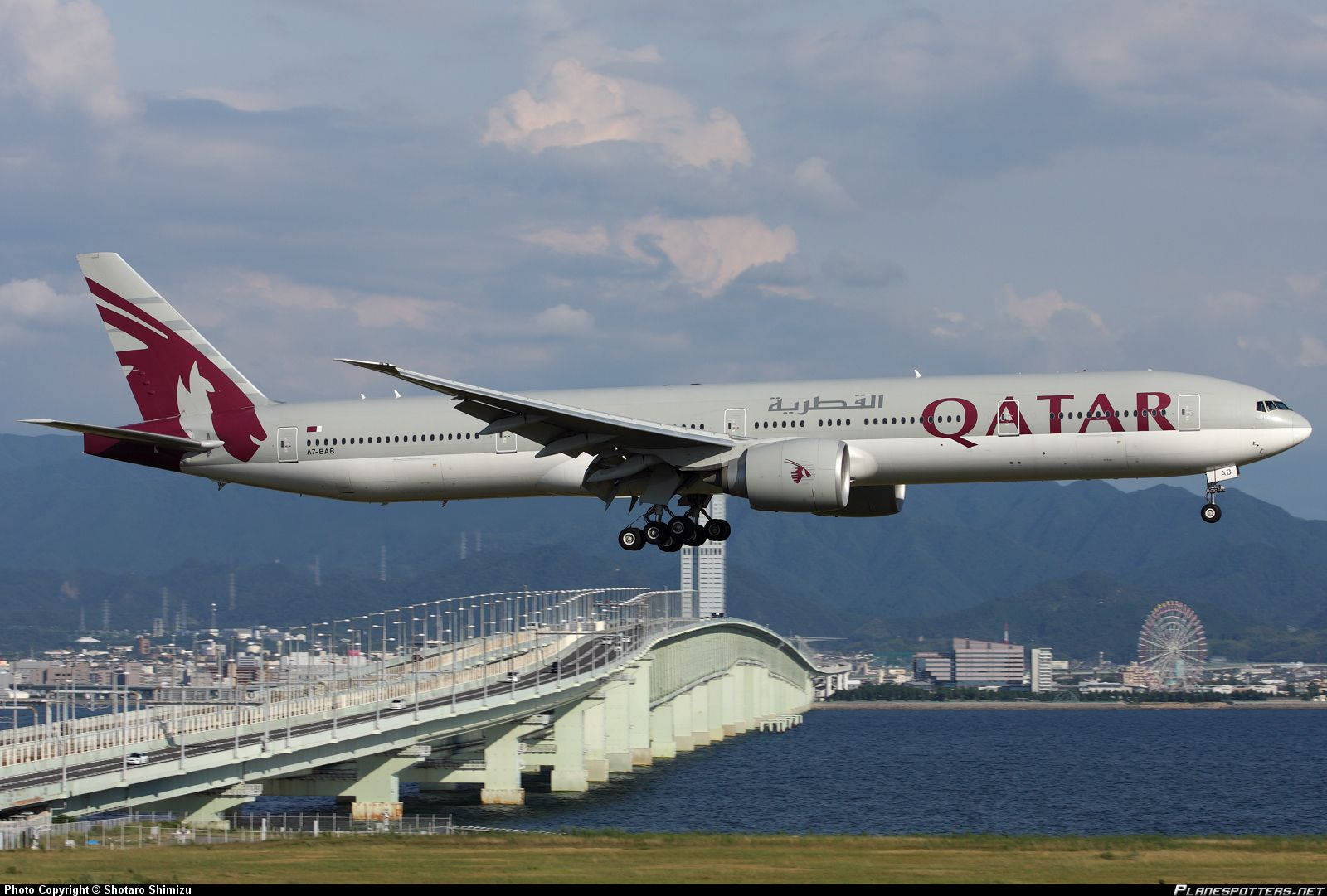 卡塔爾航空飛機起飛 壁紙 — 卡塔爾航空波音777在沿海橋樑上空低飛起飛，背景有山脈和城市景觀。