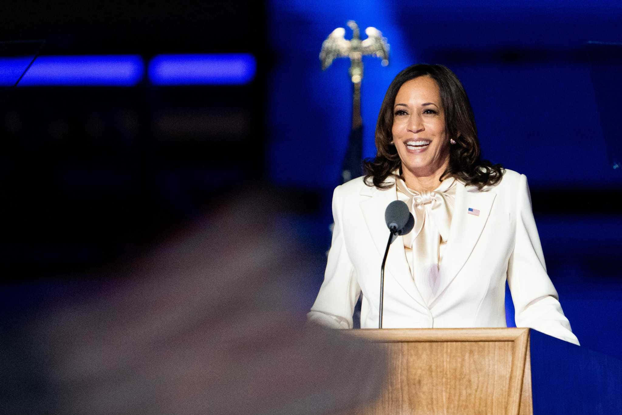 Kamala Harris 桌布 壁紙 — 卡瑪拉·哈里斯在藍色舞台燈光下，於木製講台上溫暖微笑發言，身後有鷹徽標誌。