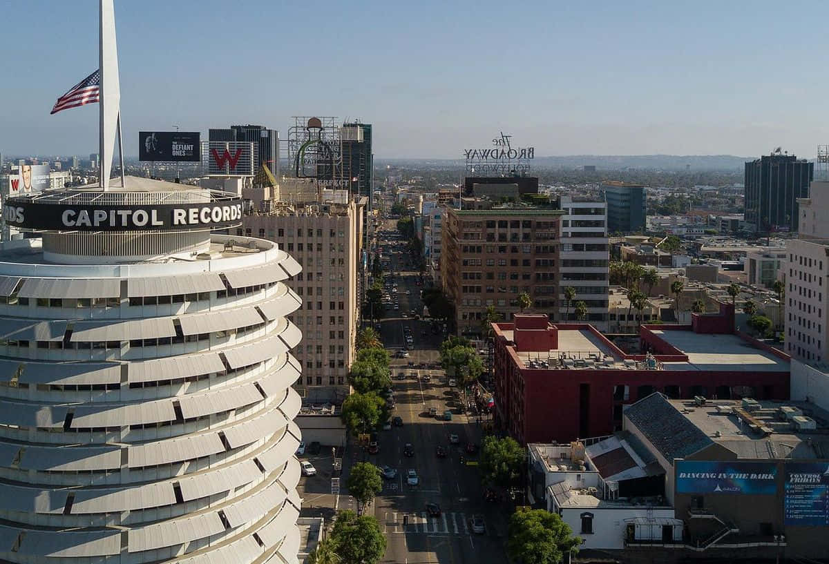 卡皮托爾唱片大樓的優秀航拍視角 壁紙 — 標誌性 Capitol Records 大樓的鳥瞰圖，頂部有美國國旗，俯瞰繁華的洛杉磯街道和周圍的城市天際線，天空清澈湛藍。
