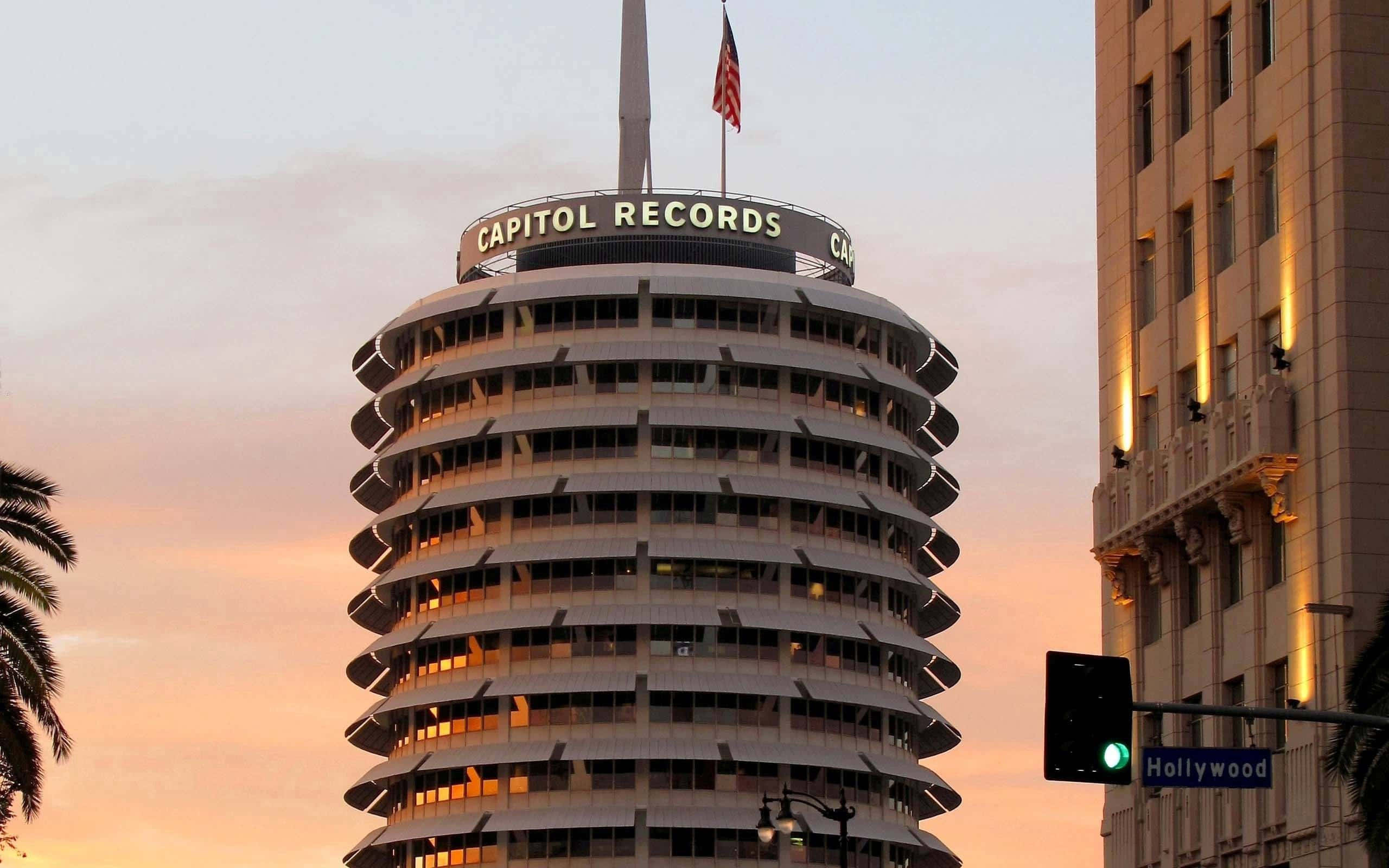 卡皮托爾唱片大樓的日落景色 壁紙 — 標誌性的 Capitol Records 大樓在日落時分閃耀著溫暖的燈光，映襯著粉紅和橙色的天空，周圍環繞著棕櫚樹和好萊塢街道標誌。