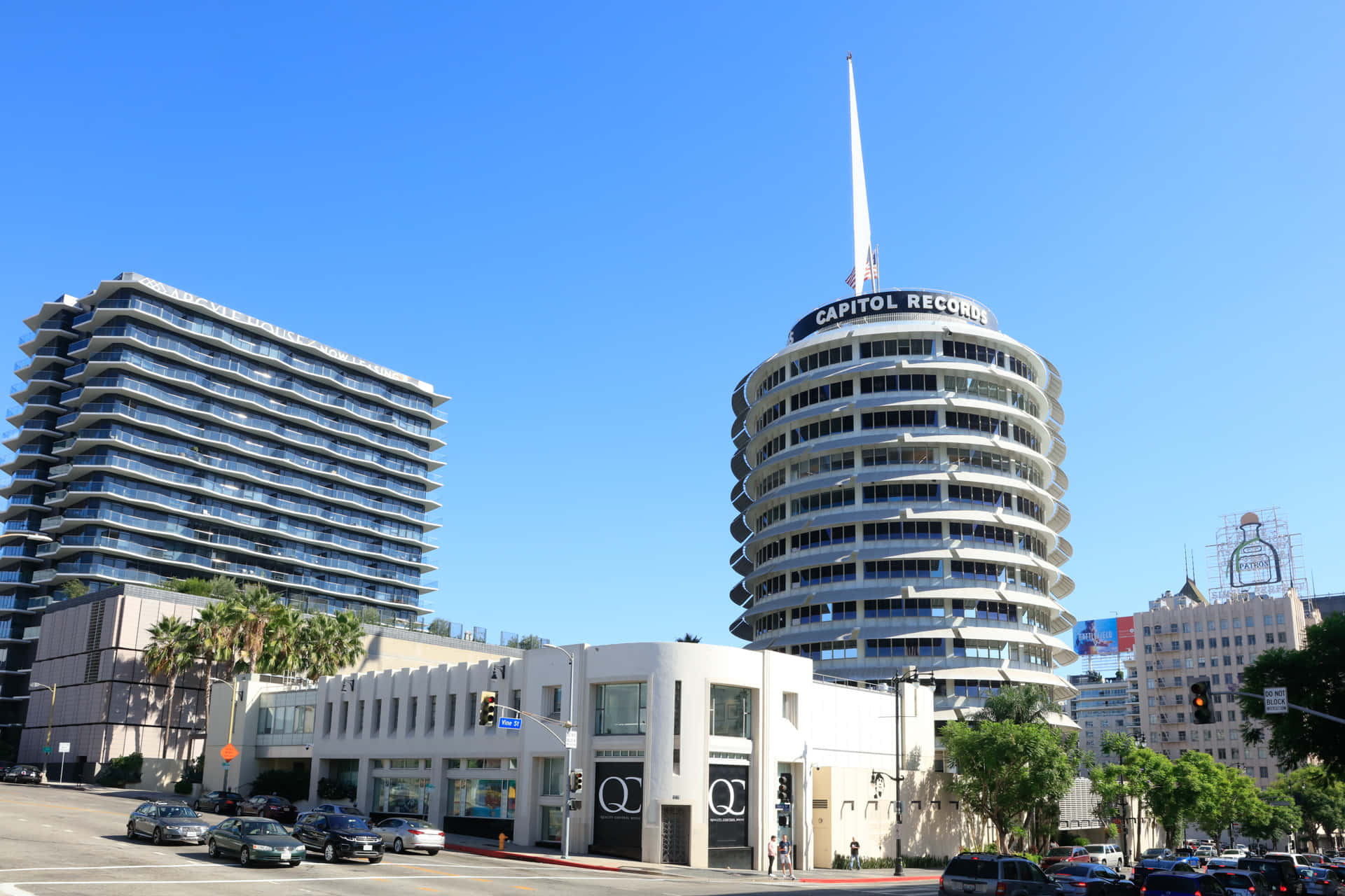 卡皮托爾唱片大樓與其他建築 壁紙 — 標誌性的 Capitol Records 大樓在晴朗的藍天下高聳矗立，現代化的玻璃塔樓和棕櫚樹排列在下方繁忙的街道兩旁。