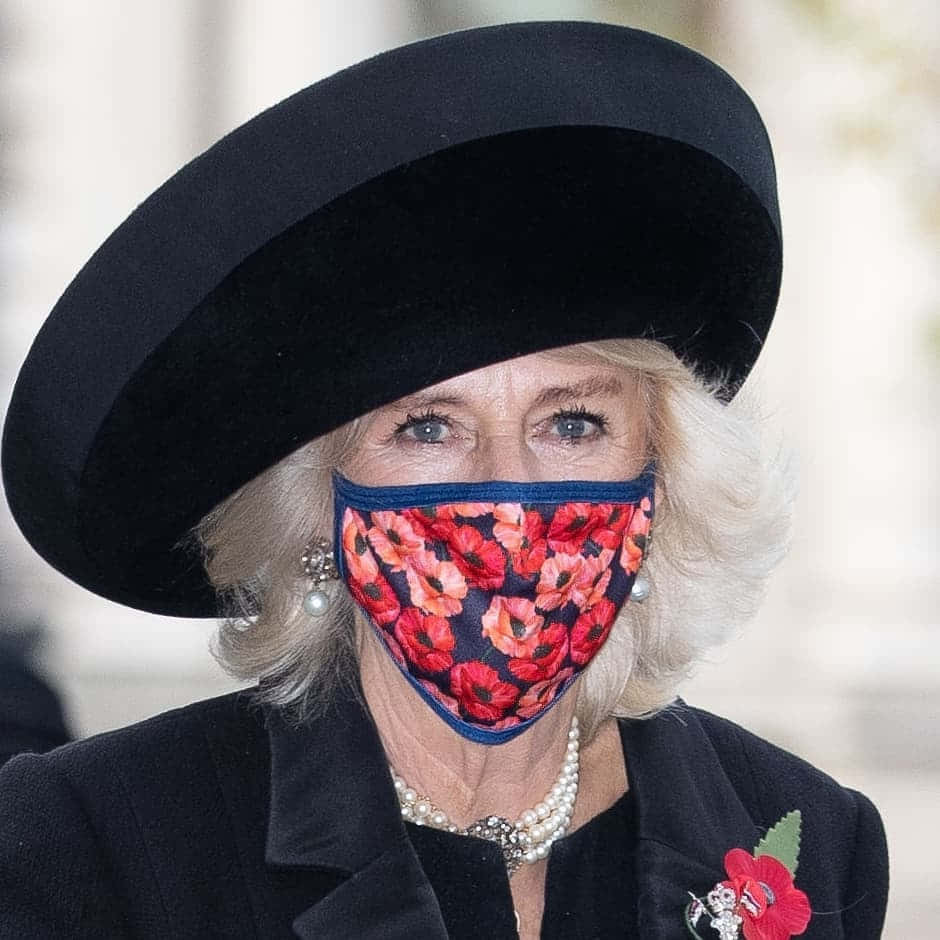 卡米拉王后佩戴虞美人面罩 壁紙 — 一位金髮女性戴著黑色帽子和紅色罌粟花圖案口罩，直視鏡頭。