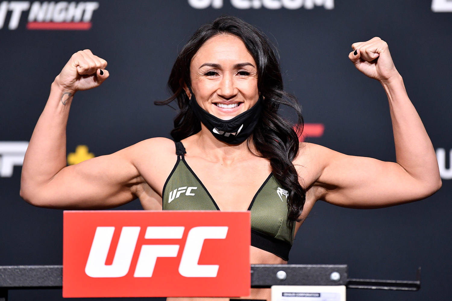 卡菈·埃斯帕薩展現肌肉 壁紙 — 一位微笑的女性UFC拳手在稱重活動中展示肌肉，穿著綠色運動胸罩和黑色口罩。