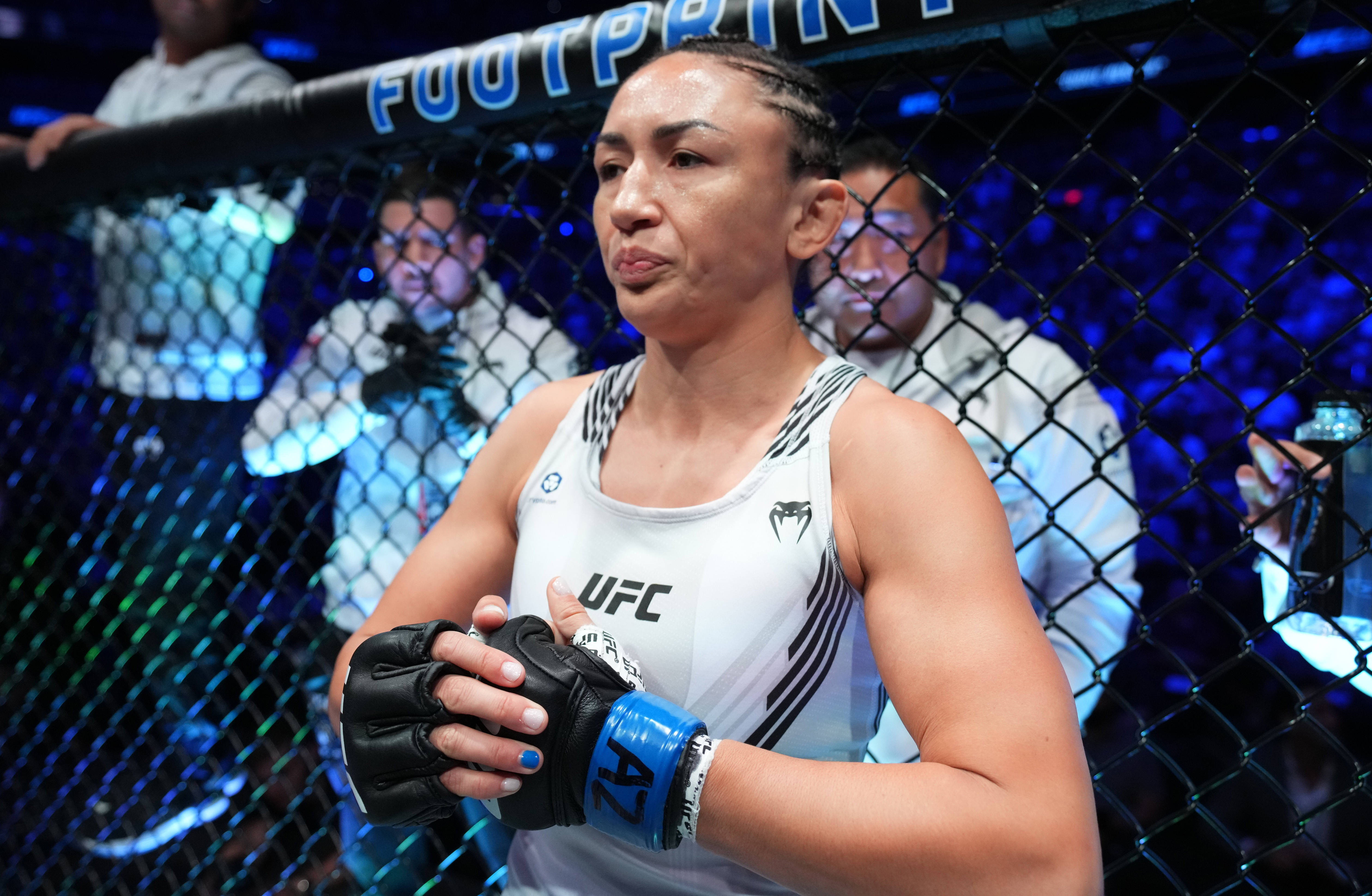 卡菈·艾斯帕薩調整拳套 壁紙 — 一位專注的女性MMA拳手身穿白色UFC背心，站在藍色燈光的八角籠中，雙手交握。