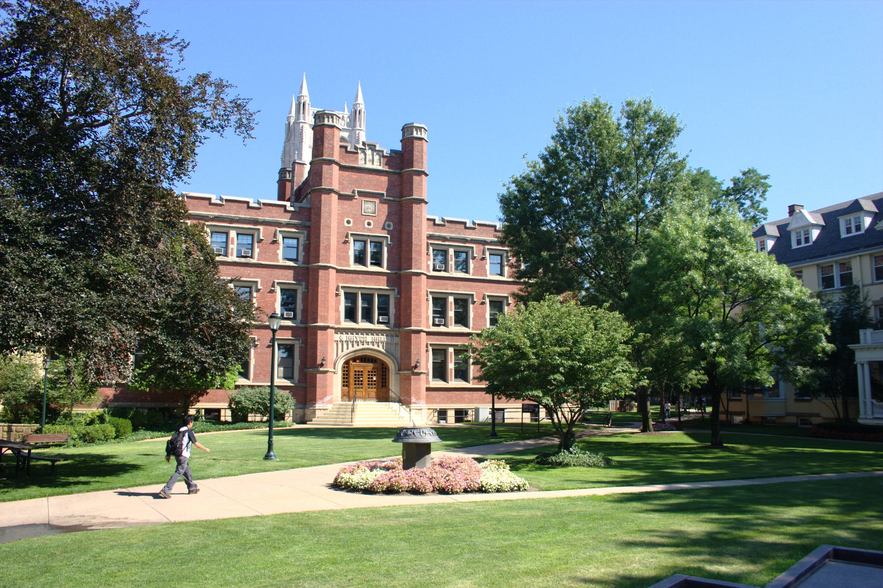 卡西西維爾大學海頓廳壯麗景觀 壁紙 — 一名學生穿過陽光普照的校園廣場，走向一座被茂密綠樹環繞的宏偉紅磚哥德式大學建築。