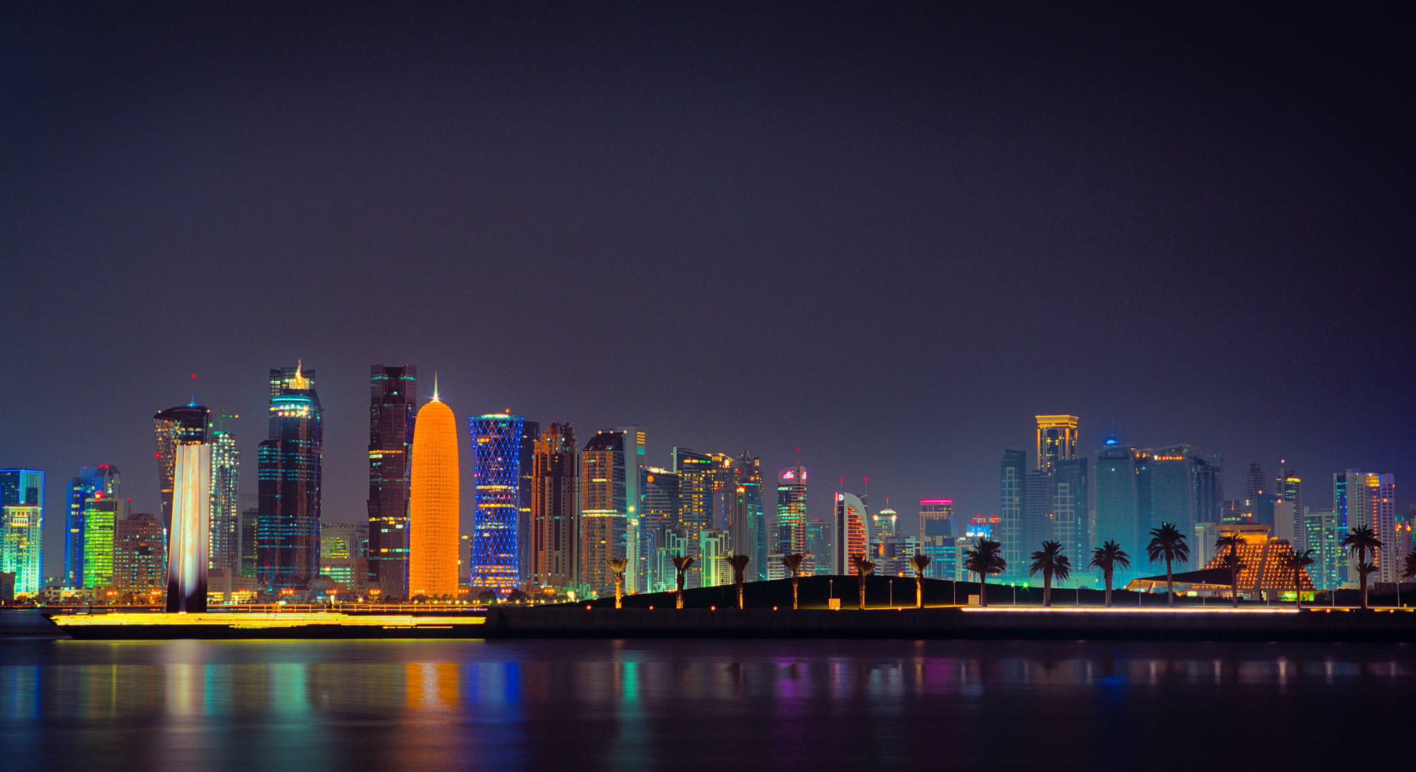 卡達創新建築 壁紙 — 令人目眩的夜景城市景觀，摩天大樓在霓虹色調中閃耀，倒映在平靜的水面上。