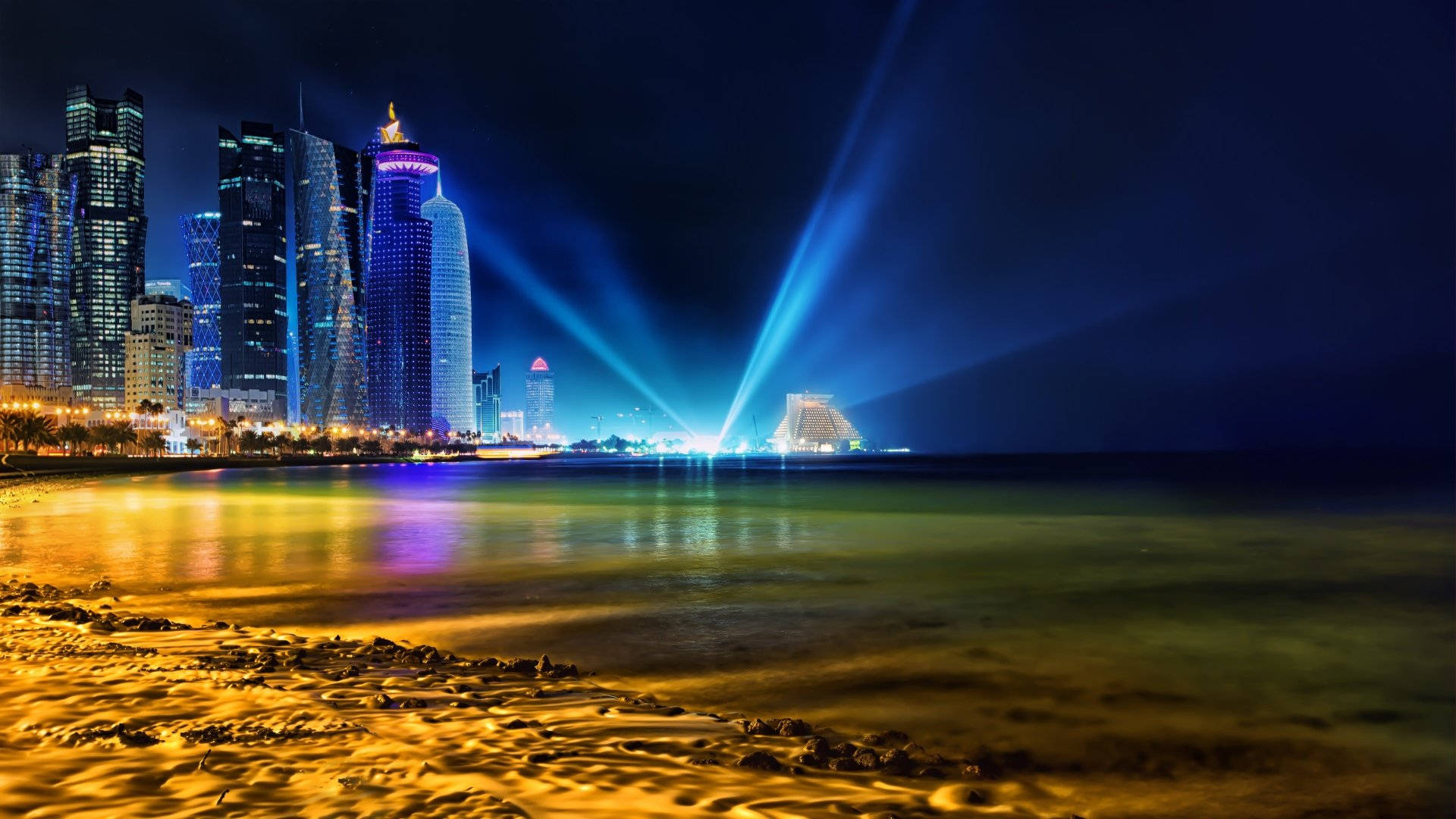 卡達海灘夜景 壁紙 — 充滿活力的夜景城市景觀，閃爍的摩天大樓和戲劇性的藍色聚光燈反射在沙灘旁的平靜水面上。