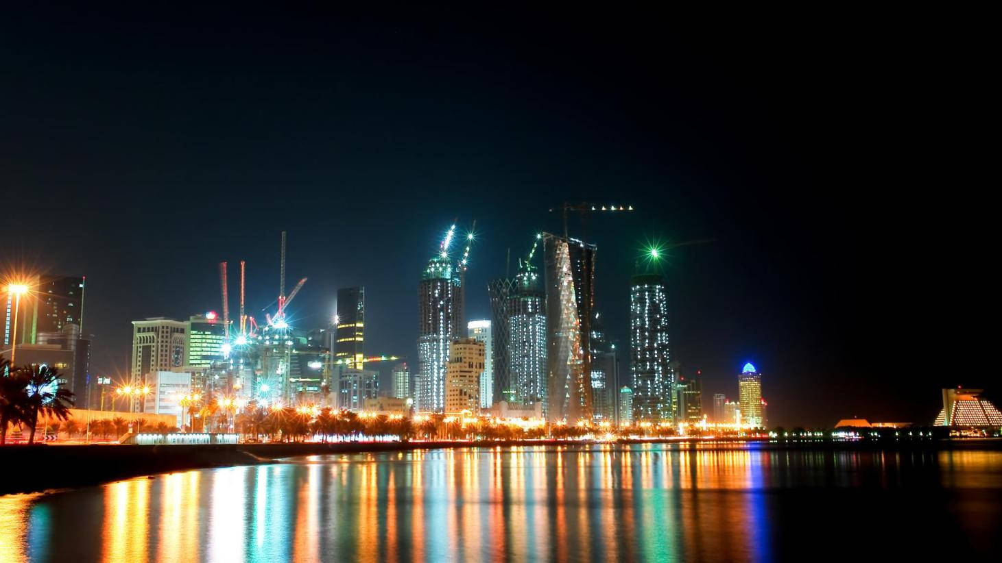 卡達璀璨建築 壁紙 — 璀璨的夜景城市景觀，燈火通明的摩天大樓和建築起重機倒映在平靜的水面上。