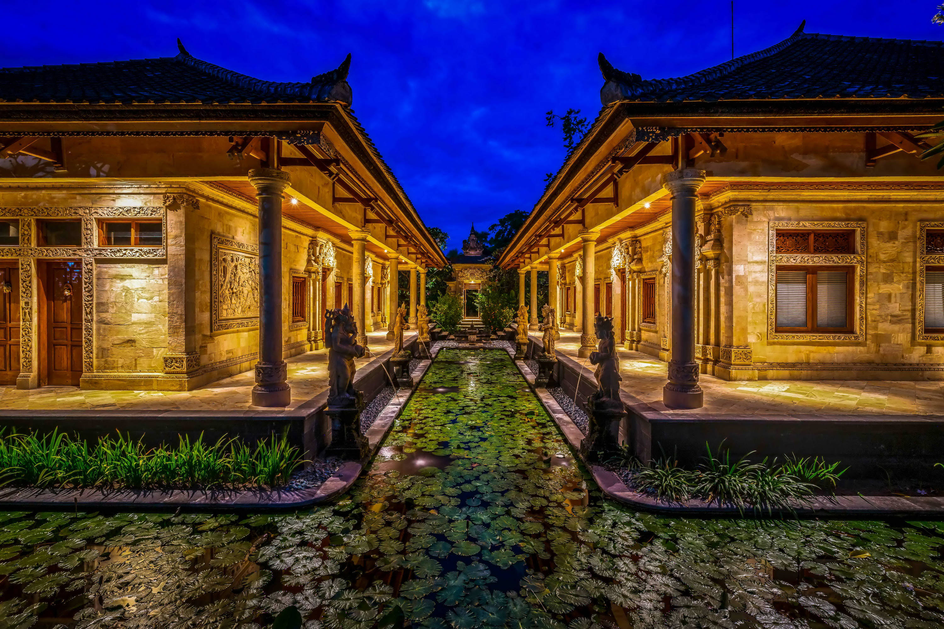 印尼馬塔哈里海灘度假村 壁紙 — 黃昏時分對稱的峇里島庭院，發光的燈籠照亮石頭建築和睡蓮池。