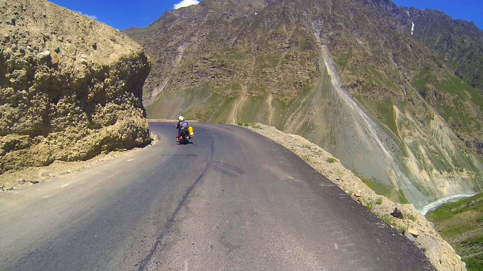 印度列城-馬納利公路摩托車離山 壁紙 — 一位孤獨的電單車手在晴朗藍天下，沿著蜿蜒的山路行駛，周圍是崎嶇的山峰和稀疏的綠意。