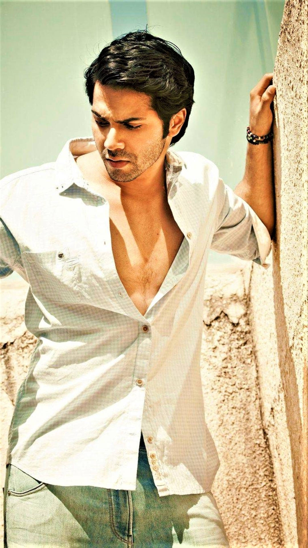 印度型男 Varun Dhawan 壁紙 — 一名身穿白恤衫的男子在明亮的陽光下靠著紋理牆壁，表情嚴肅。