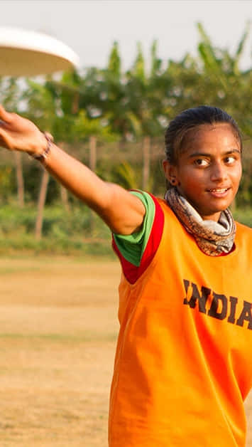 印度女性運動員於 Android Ultimate 飛盤背景 壁紙 — 一位身穿橙色「INDIA」球衣的年輕女子微笑著在戶外擲飛盤，背景是綠樹和乾草。