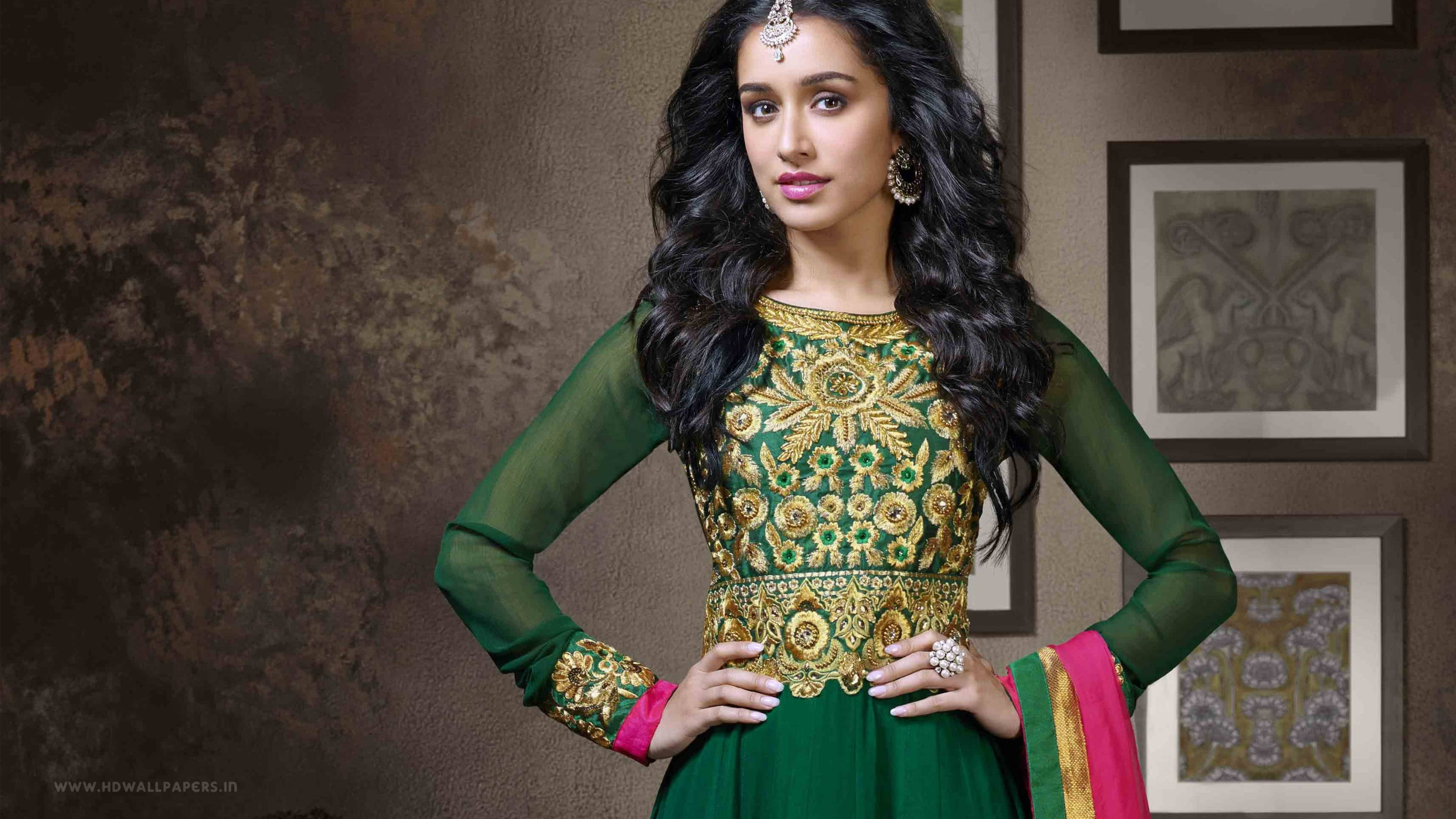 印度女星Shraddha Kapoor身著亮片裝束展現迷人風采 壁紙 — 一位身穿綠色和金色刺繡傳統服裝的女性，自信地在帶有紋理牆壁和裱框藝術品前擺姿勢。