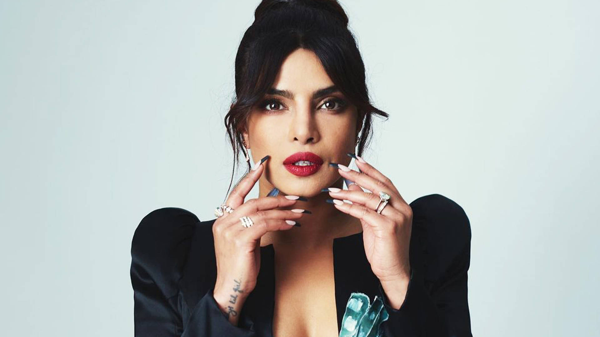 印度女演員普里揚卡·喬普拉驚訝表情 壁紙 — Priyanka Chopra 自信地擺姿勢，雙手靠近臉部，背景為純淺灰色，身穿黑色西裝外套和紅色唇膏。