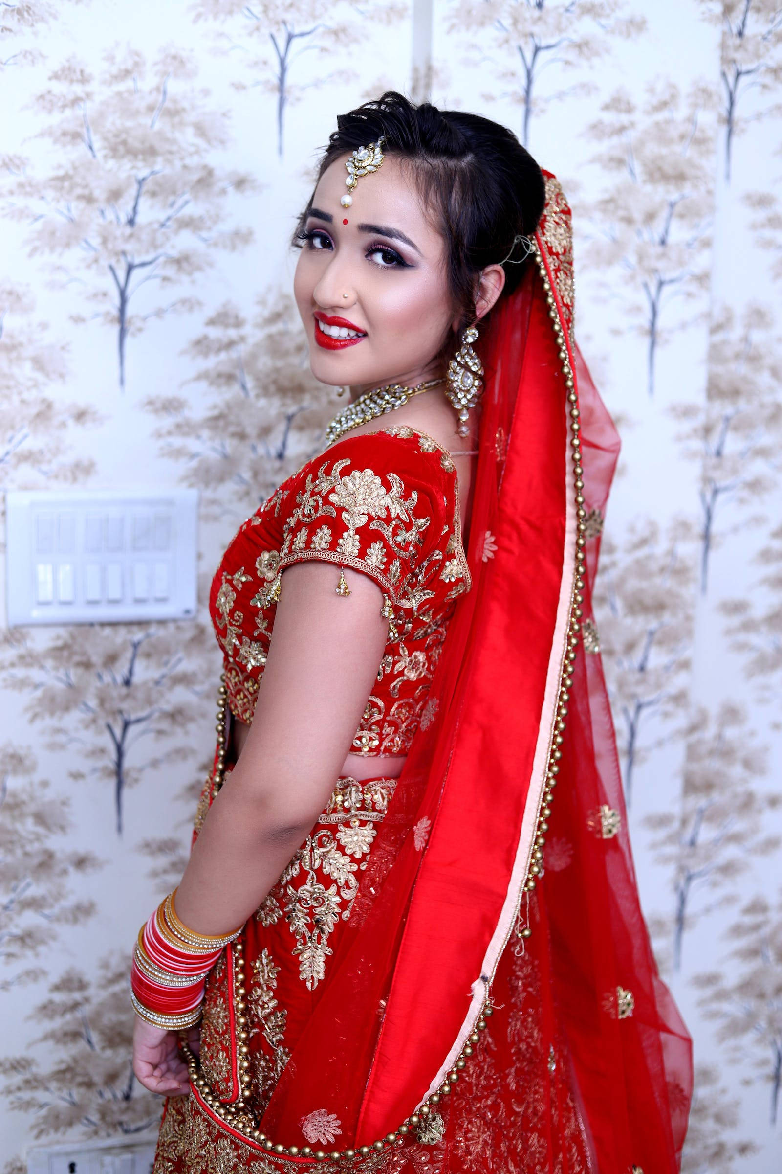 印度婚禮紅金紗 壁紙 — 一位身穿紅金刺繡新娘禮服（lehenga）的耀眼女性，在樹紋壁紙前優雅擺姿。