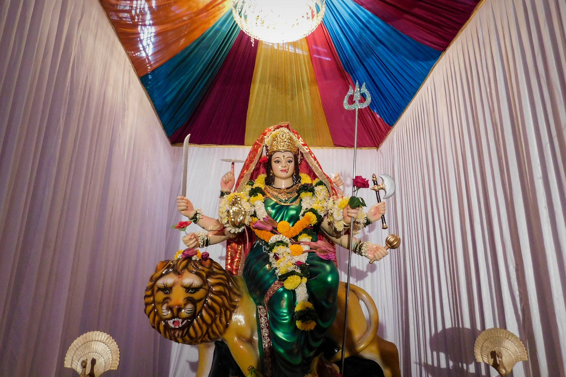 印度教女神杜爾加彩色窗簾 壁紙 — 一尊色彩鮮豔的多臂女神雕像，身穿綠色服飾，騎著獅子，在色彩繽紛的垂簾天篷下，飾有鮮花和三叉戟。
