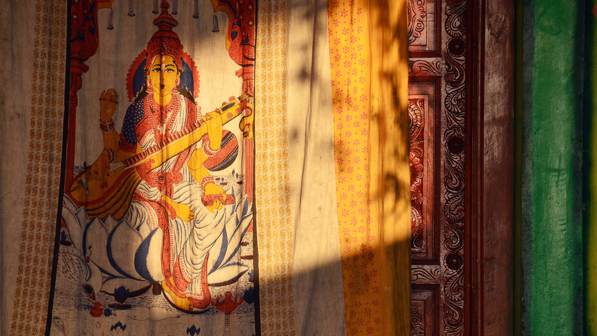 印度教女神薩拉斯瓦蒂壁毯 壁紙 — 一幅充滿活力的傳統印度神祇畫作，沐浴在透過圖案窗簾過濾的金色陽光中。