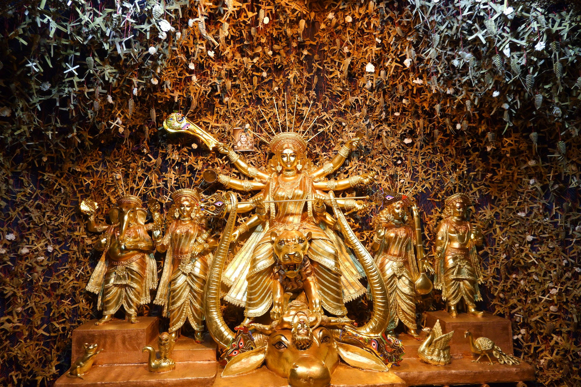印度教女神金身雕像 壁紙 — 金色多臂多武器的杜爾迦雕像，周圍環繞著較小的金色神祇，背景是懸掛的紙鶴。