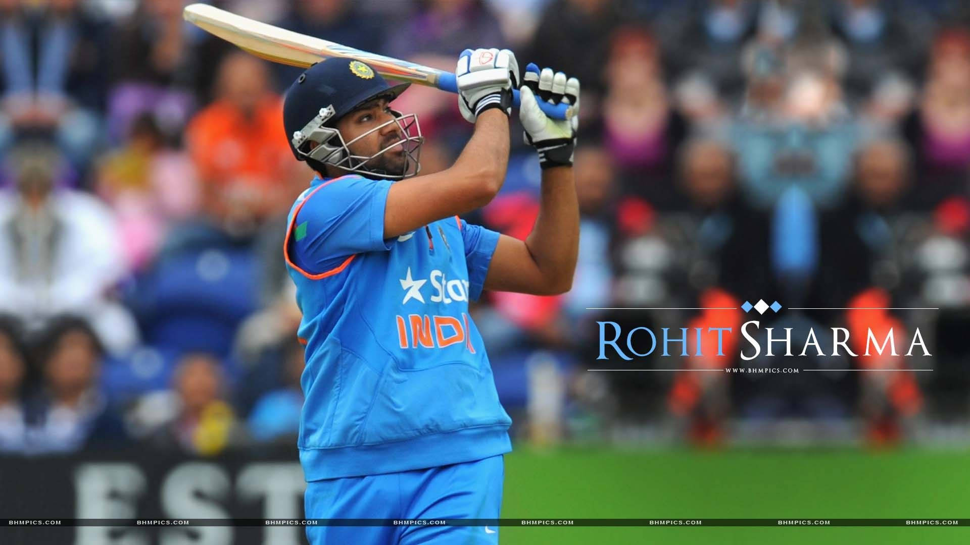 印度板球員羅希特·夏爾馬 壁紙 — 板球運動員 Rohit Sharma 身穿藍色印度球衣，在揮棒中途舉起球棒，背景是模糊的體育場人群。