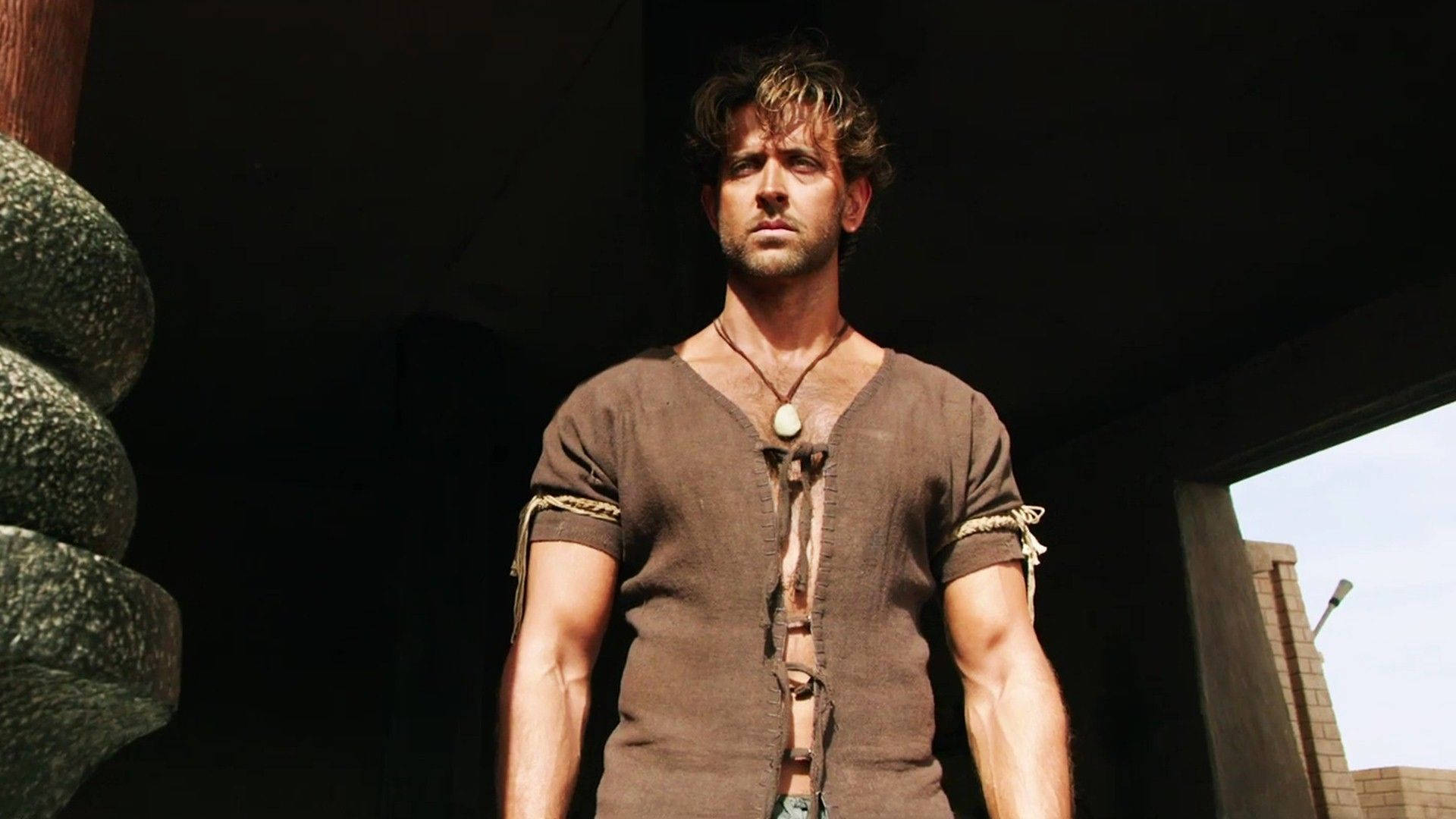 一個強壯的男人穿著棕色長袍，站在石門中，陽光照射在他的臉上。