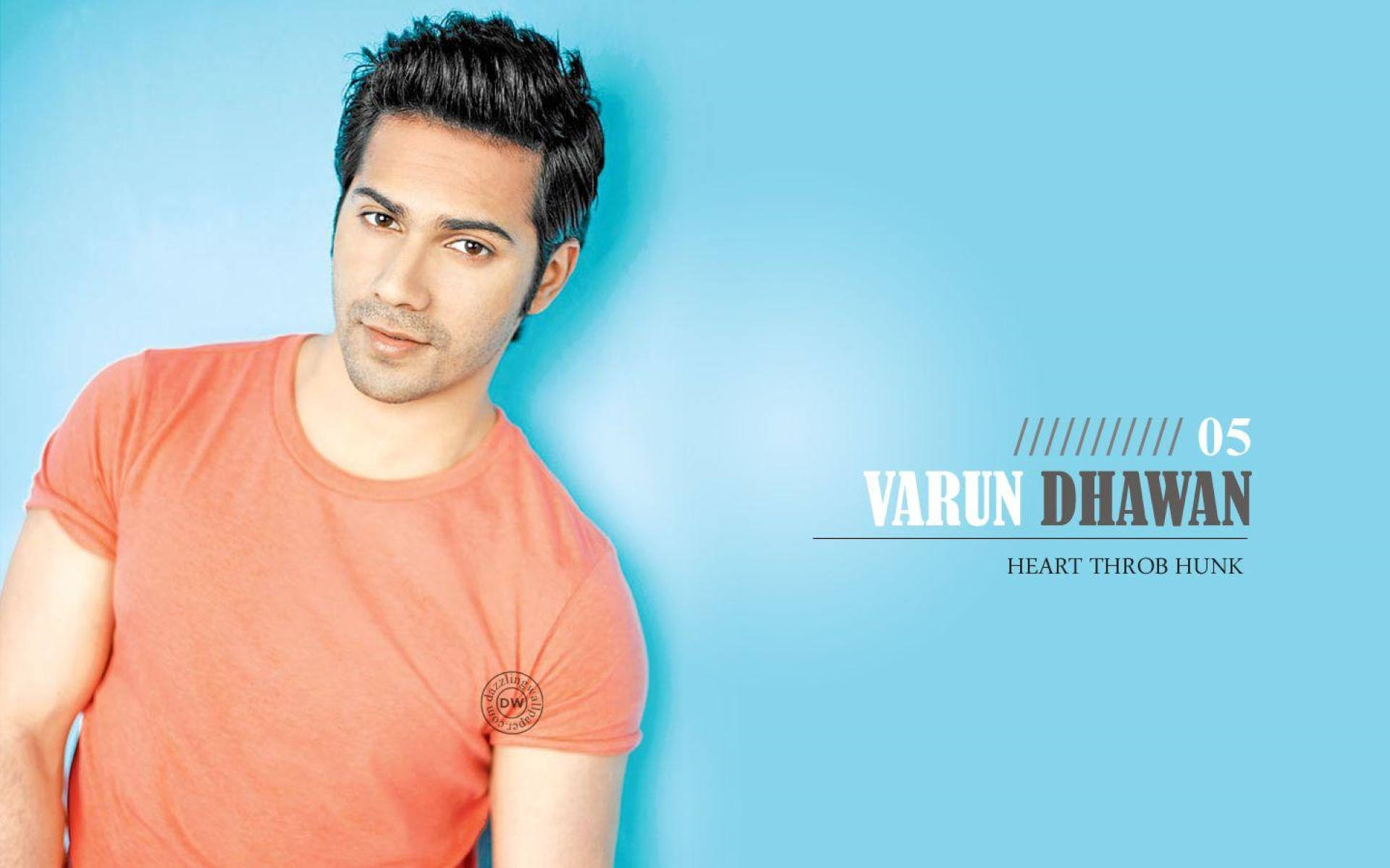 印度男神 Varun Dhawan 壁紙 — 一名身穿橙色T恤的男子在純藍色背景前擺姿勢，並帶有文字疊加。