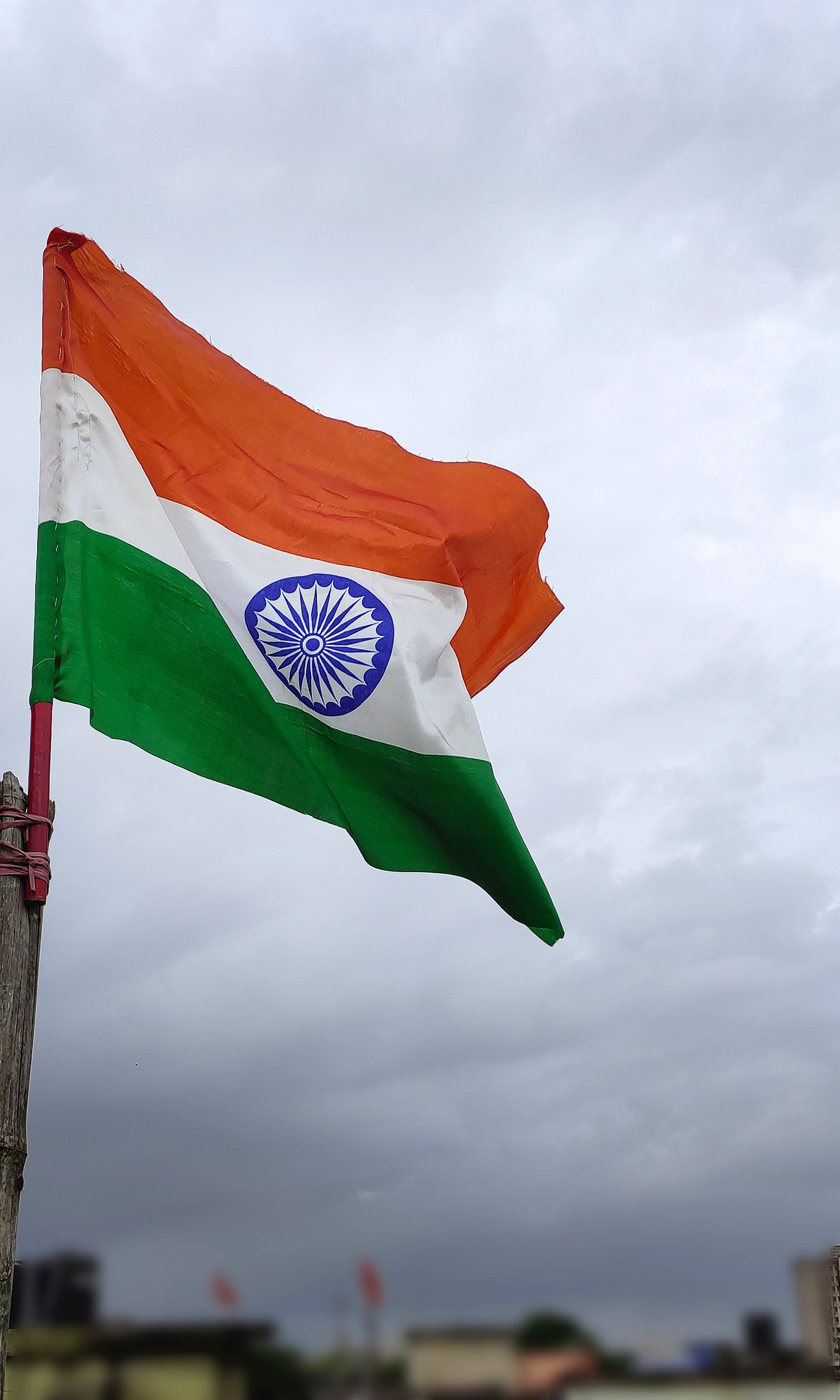 Indian 桌布 壁紙 — 印度的三色旗在陰沉多雲的天空下自豪地飄揚，遠處的城市建築模糊不清。