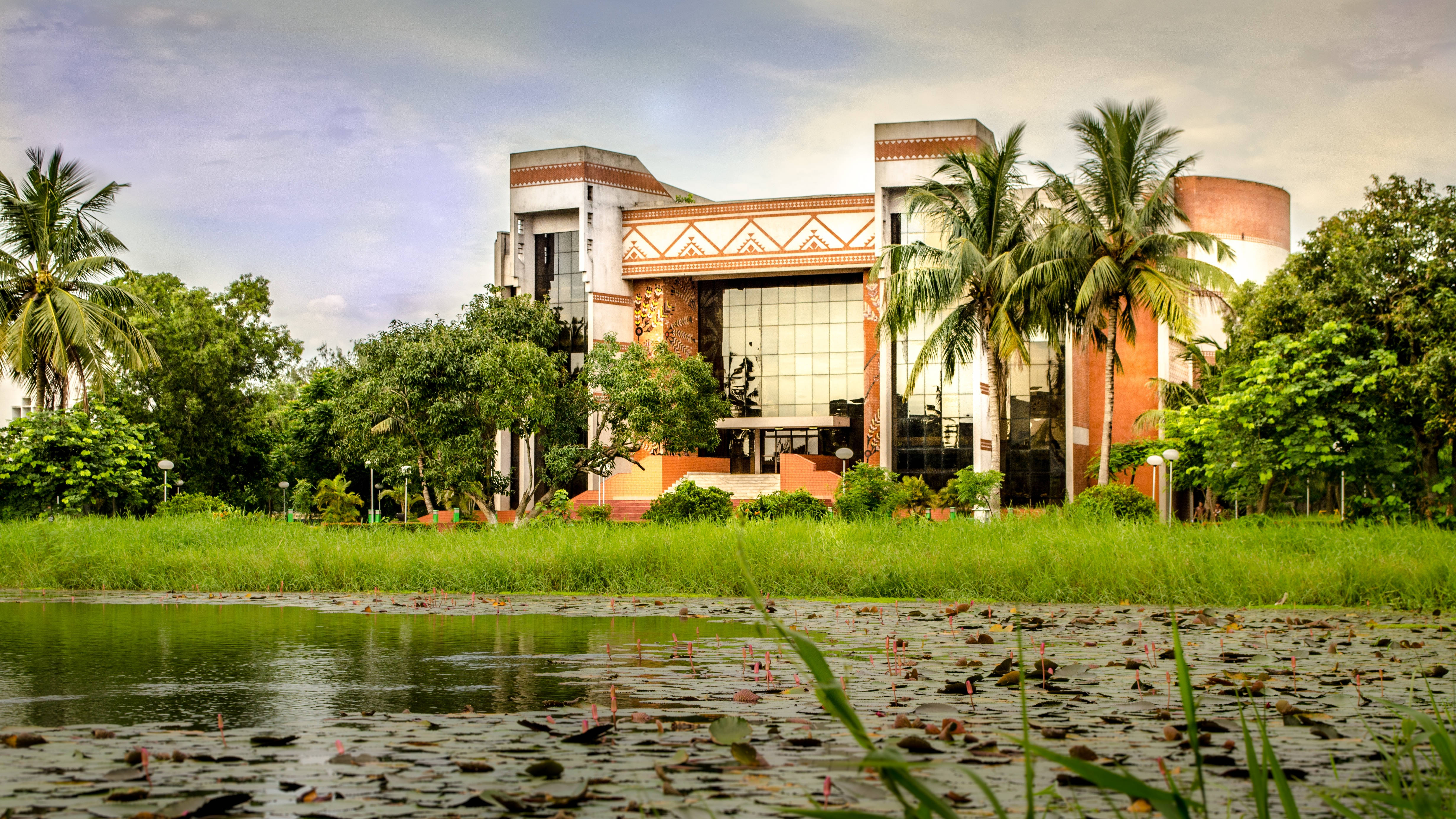 印度管理學院 Kolkata 演講廳 壁紙 — 一座現代建築，帶有陶土色調和玻璃窗，矗立在茂密的綠草和點綴著睡蓮的池塘後面，天空柔和。