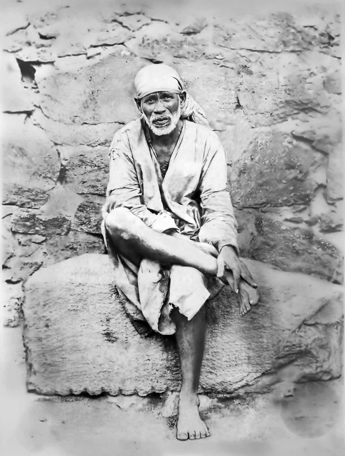 印度聖人賽巴巴（Shirdi）灰階畫像 壁紙 — 一位留著白鬍子、戴頭巾的長者赤腳坐在石壁前的石台上，這是一張復古黑白照片。