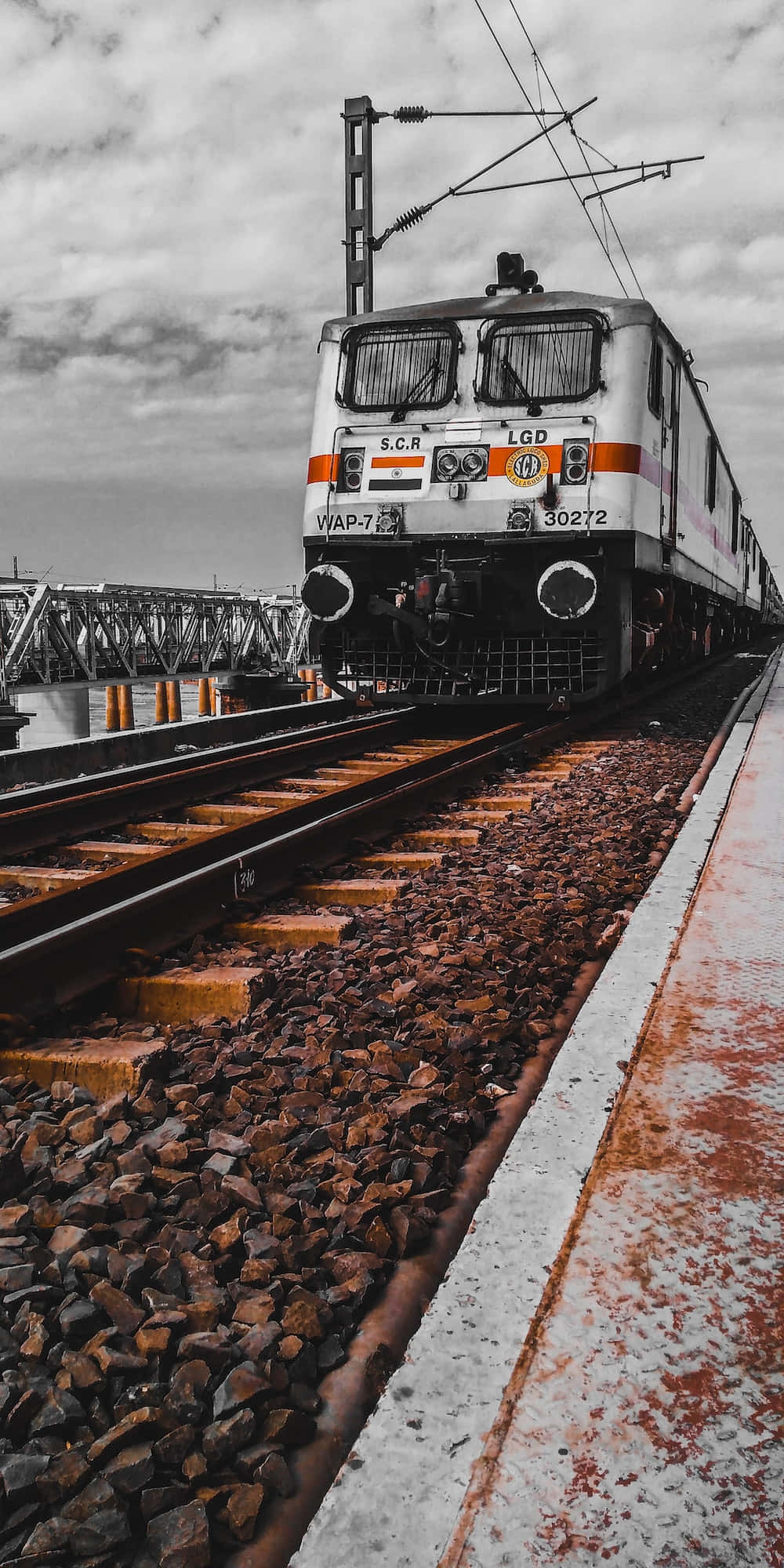 印度鐵路風景 壁紙 — 一輛帶有橙色條紋的白色印度電力機車，在陰天之下沿著生鏽的軌道靠近月台。